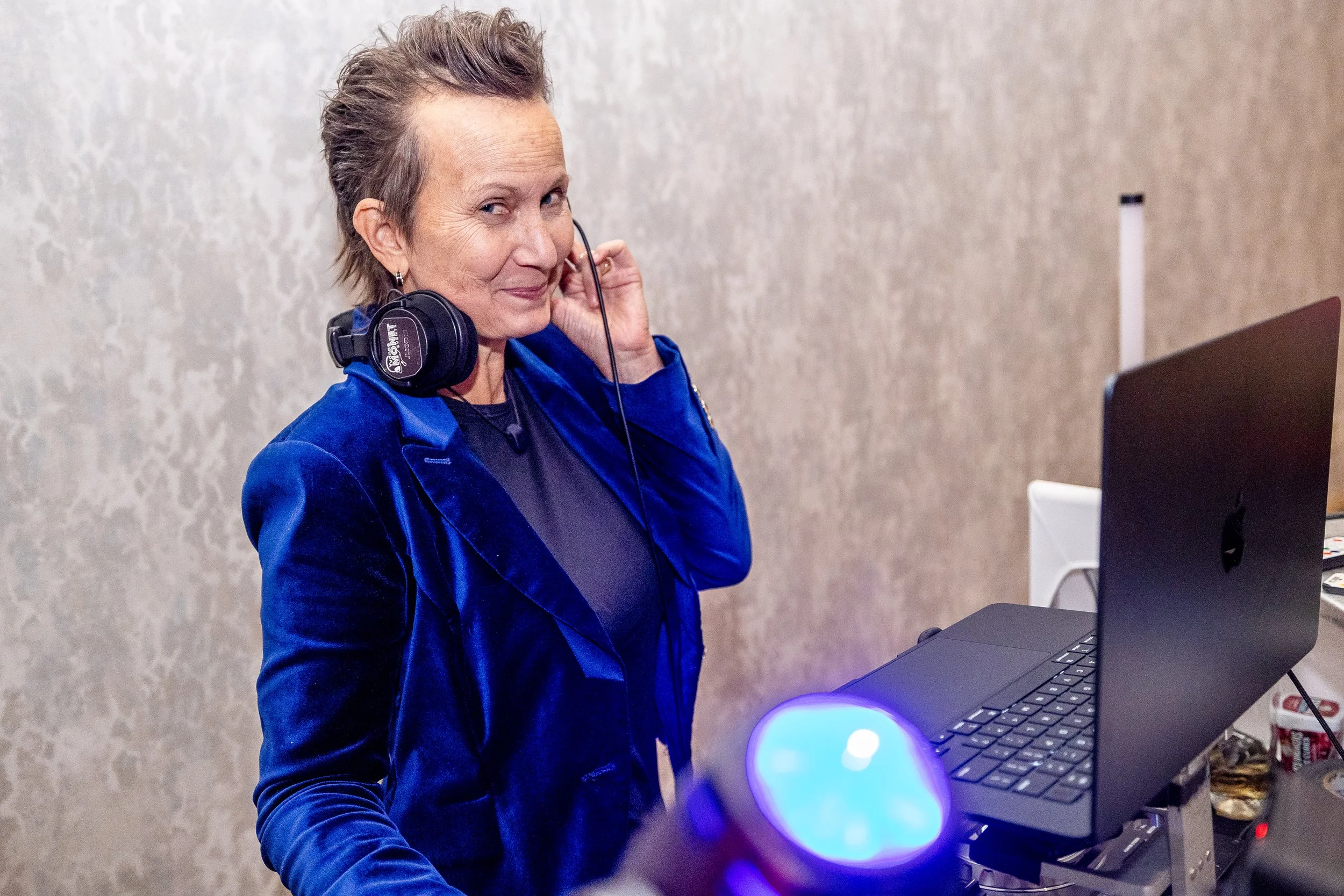 A woman wearing a blue velvet blazer and black shirt, with headphones around her neck, standing next to a laptop and smiling at the camera.