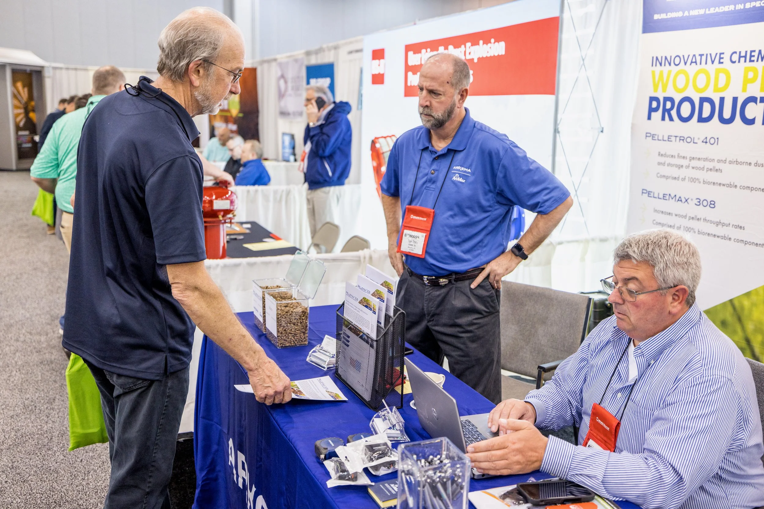 A man with gray hair and glasses is speaking to a representative at a trade show booth. The booth has informational materials and displays about wood pellet products. The representative is seated at a table with a laptop and various items, while anot