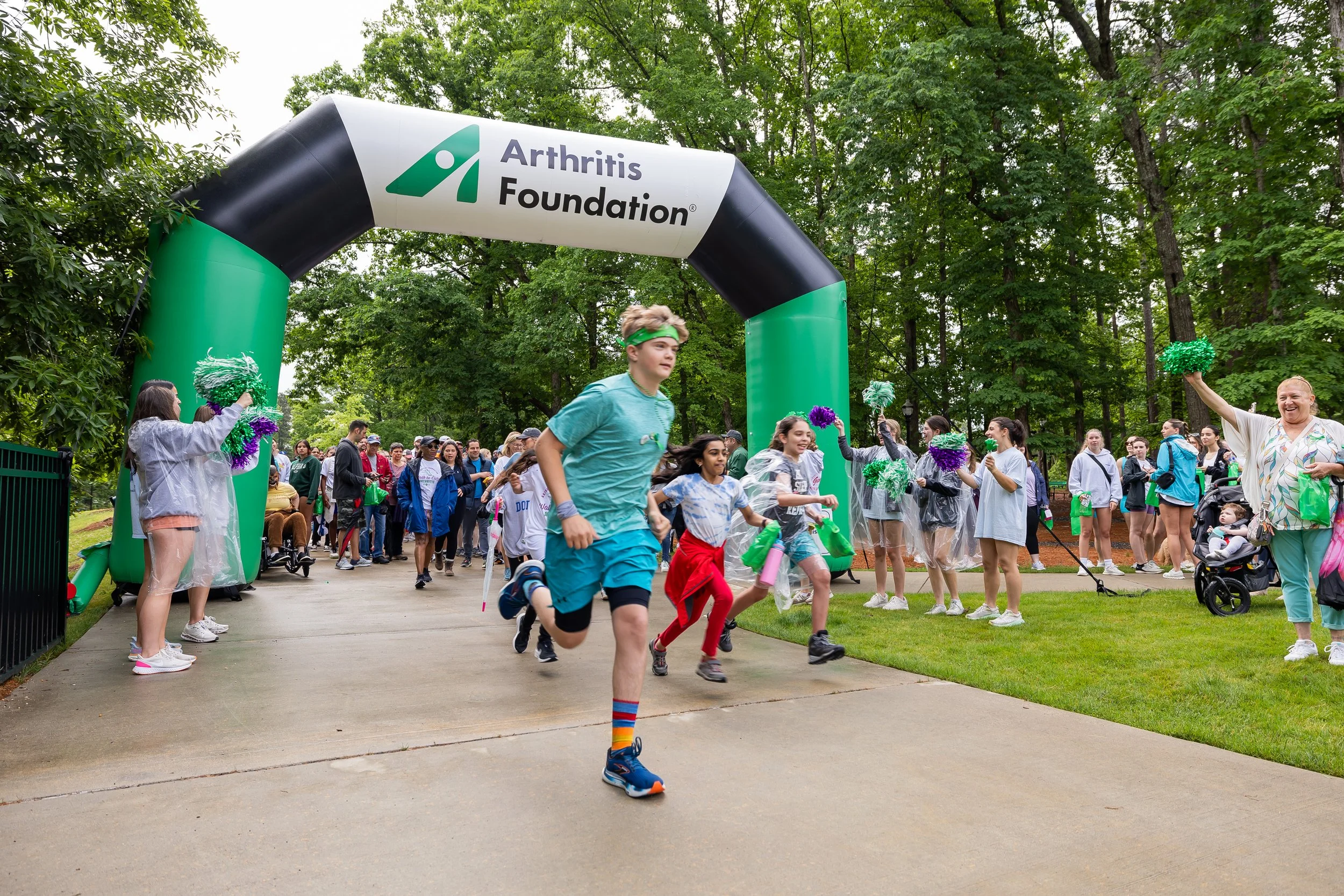 Children and adults running through an outdoor park archway for an event supporting the Arthritis Foundation, with cheerleaders on each side cheering them on.