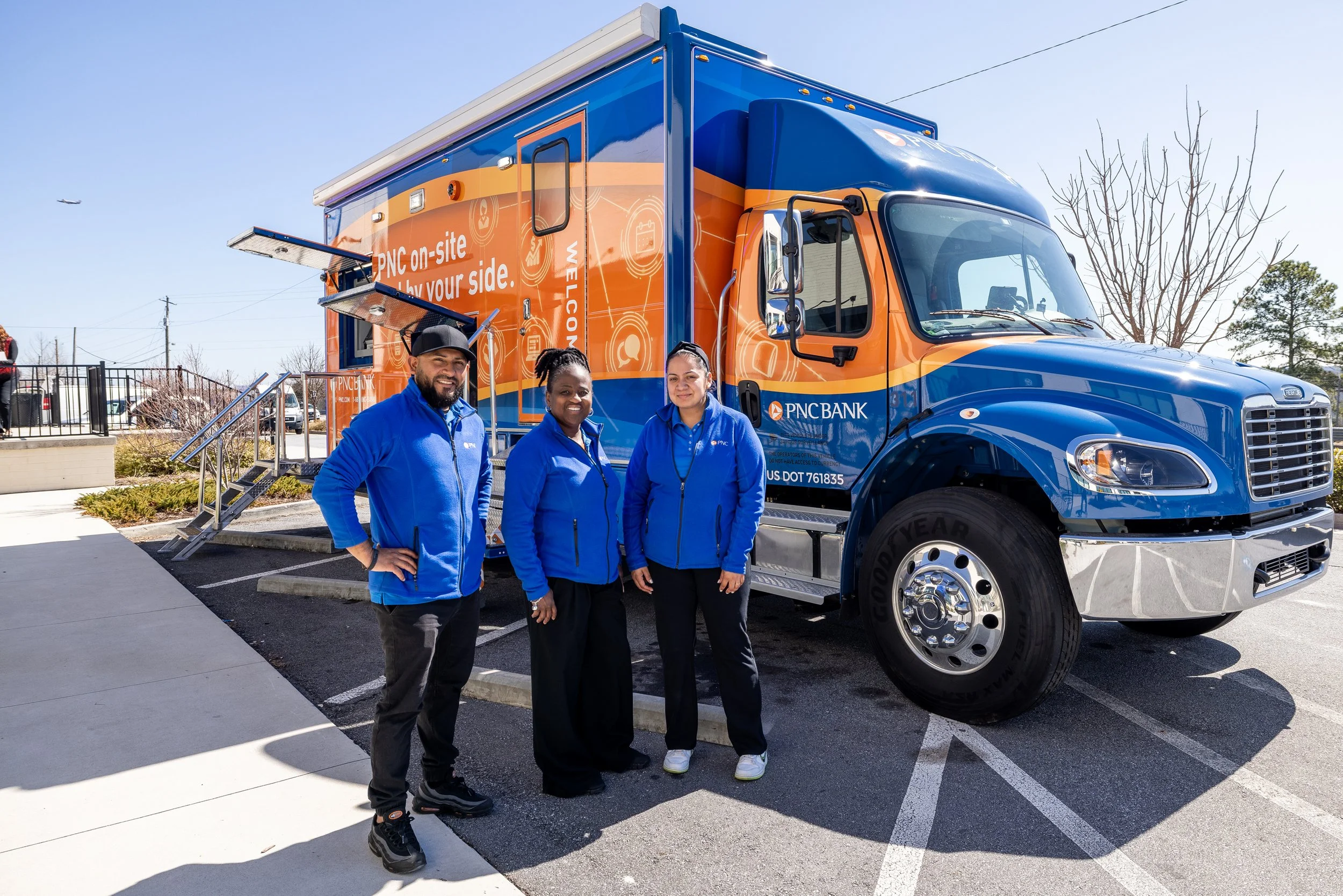 Three people standing in front of a blue and orange PNC Bank promotional mobile unit in a parking lot on a clear day.