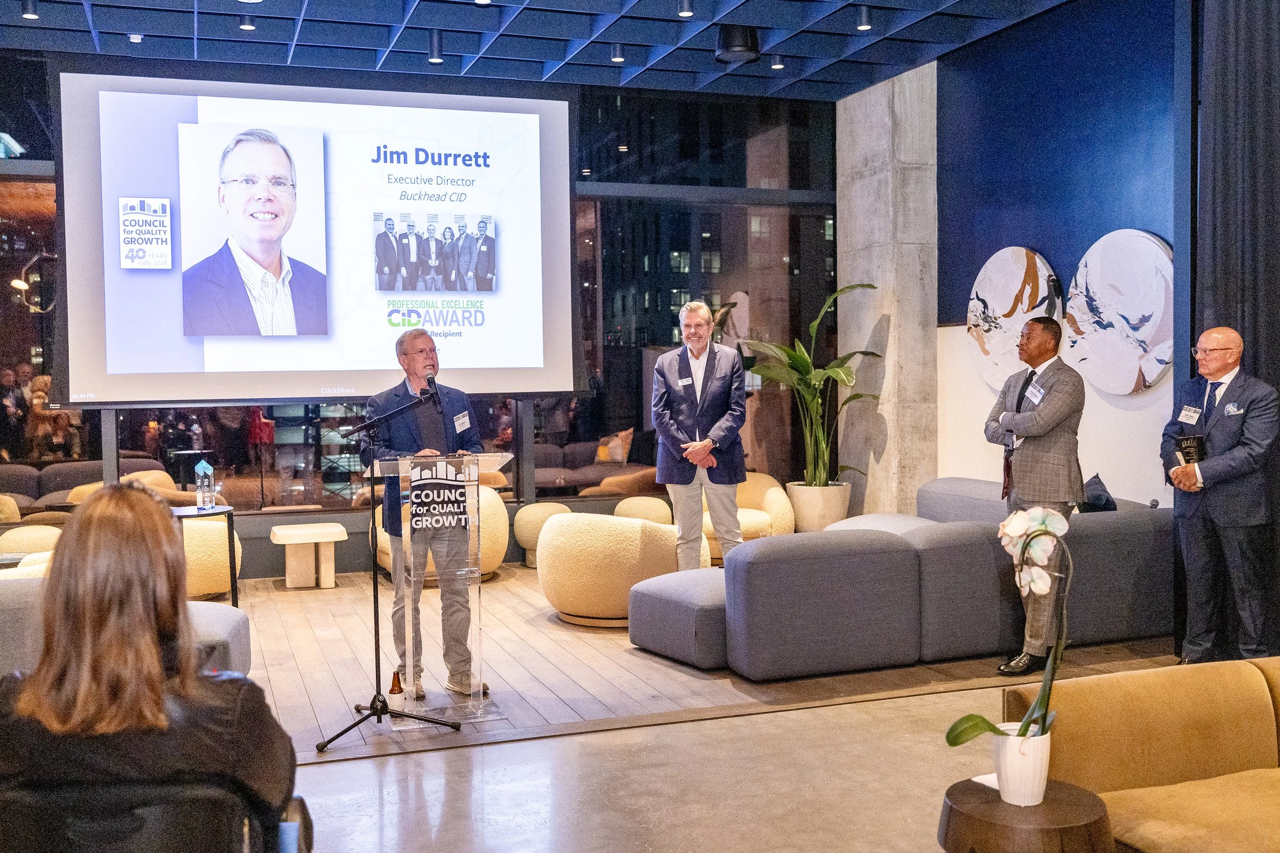 A man at a podium giving a speech during an award ceremony, with a large screen behind displaying his photo, name, and title. Four men stand to the right, one in a plaid suit with arms crossed, another with glasses holding a book, and one in a blazer
