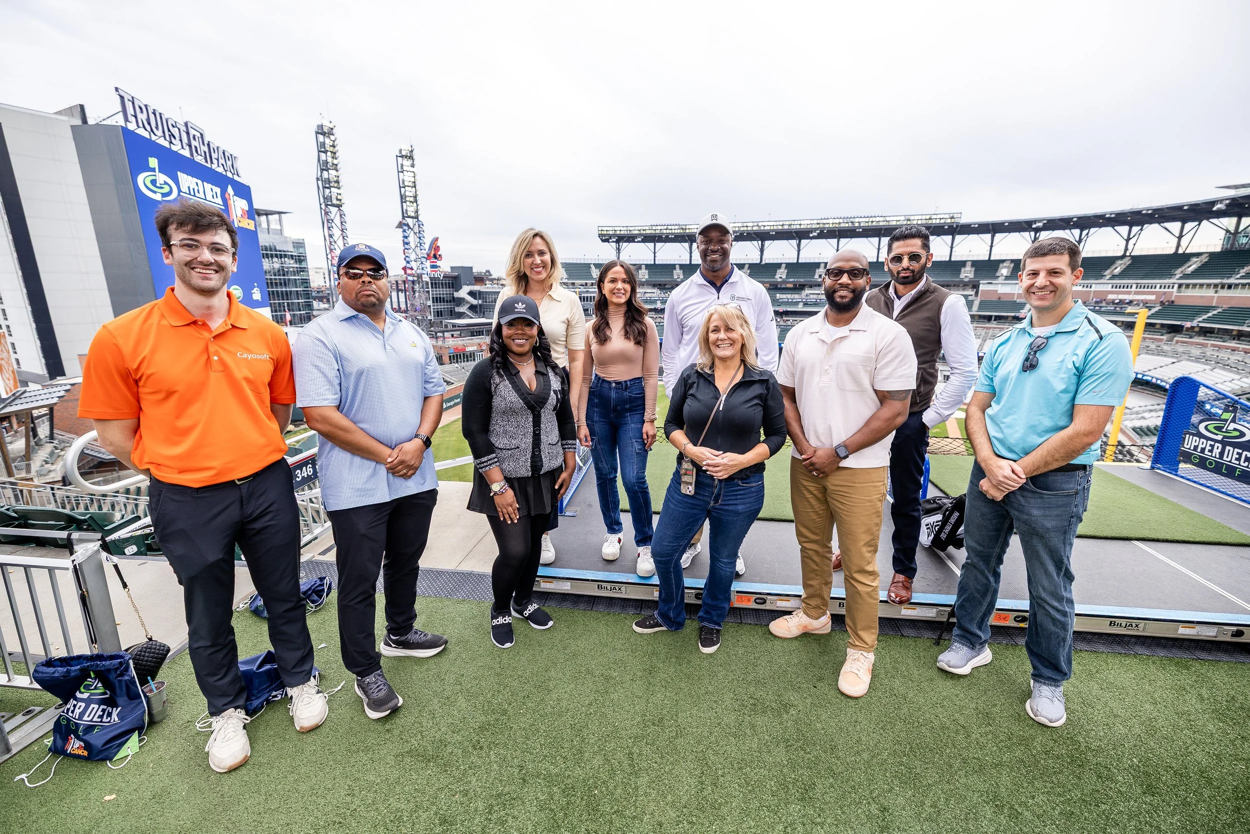 Group of diverse people standing on a platform at a sports stadium, posing for a photo with empty stadium seats in the background.