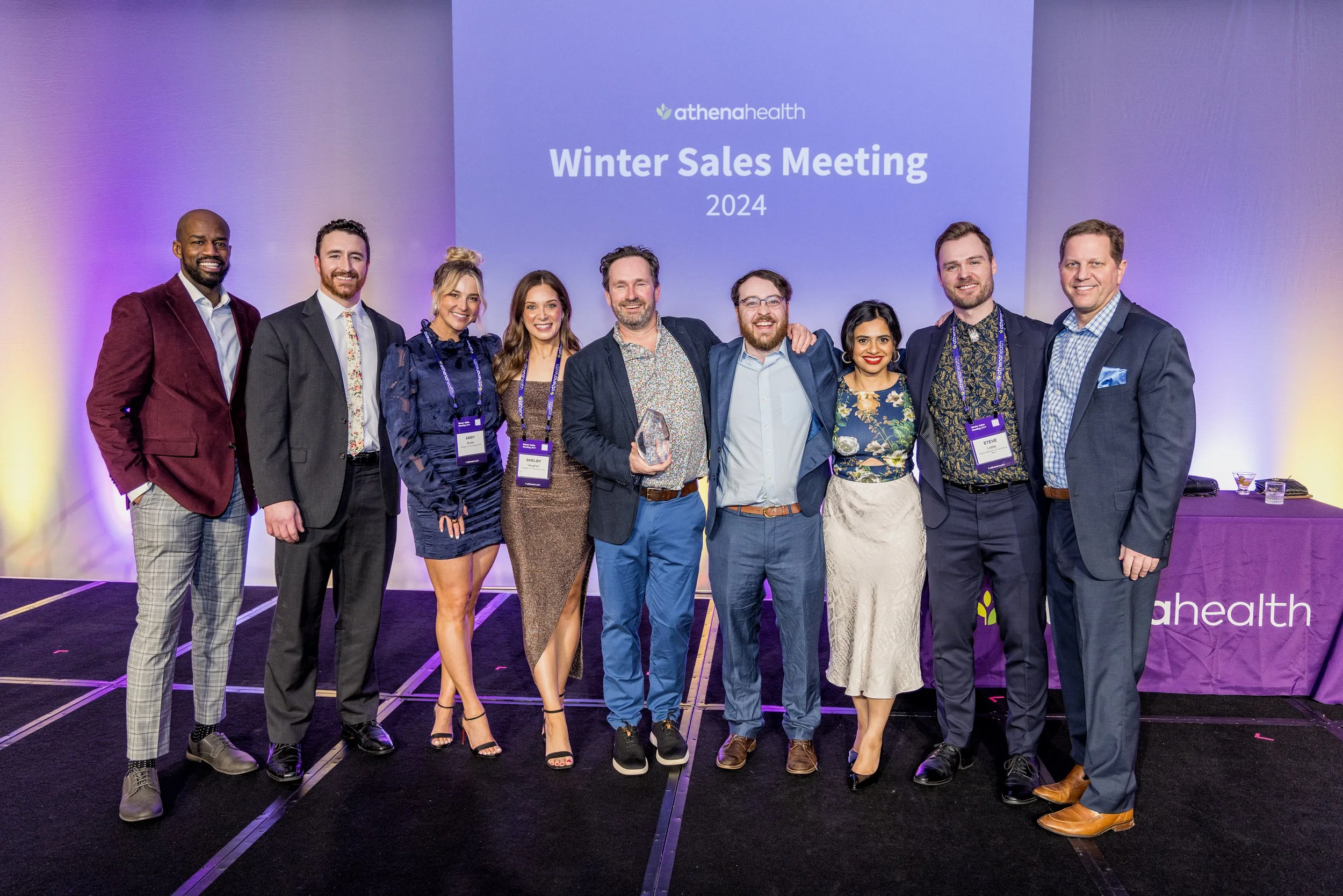 A group of ten diverse professionals standing on stage at AthenaHealth's Winter Sales Meeting 2024, posing for a photo, with a large purple screen behind them displaying event details.