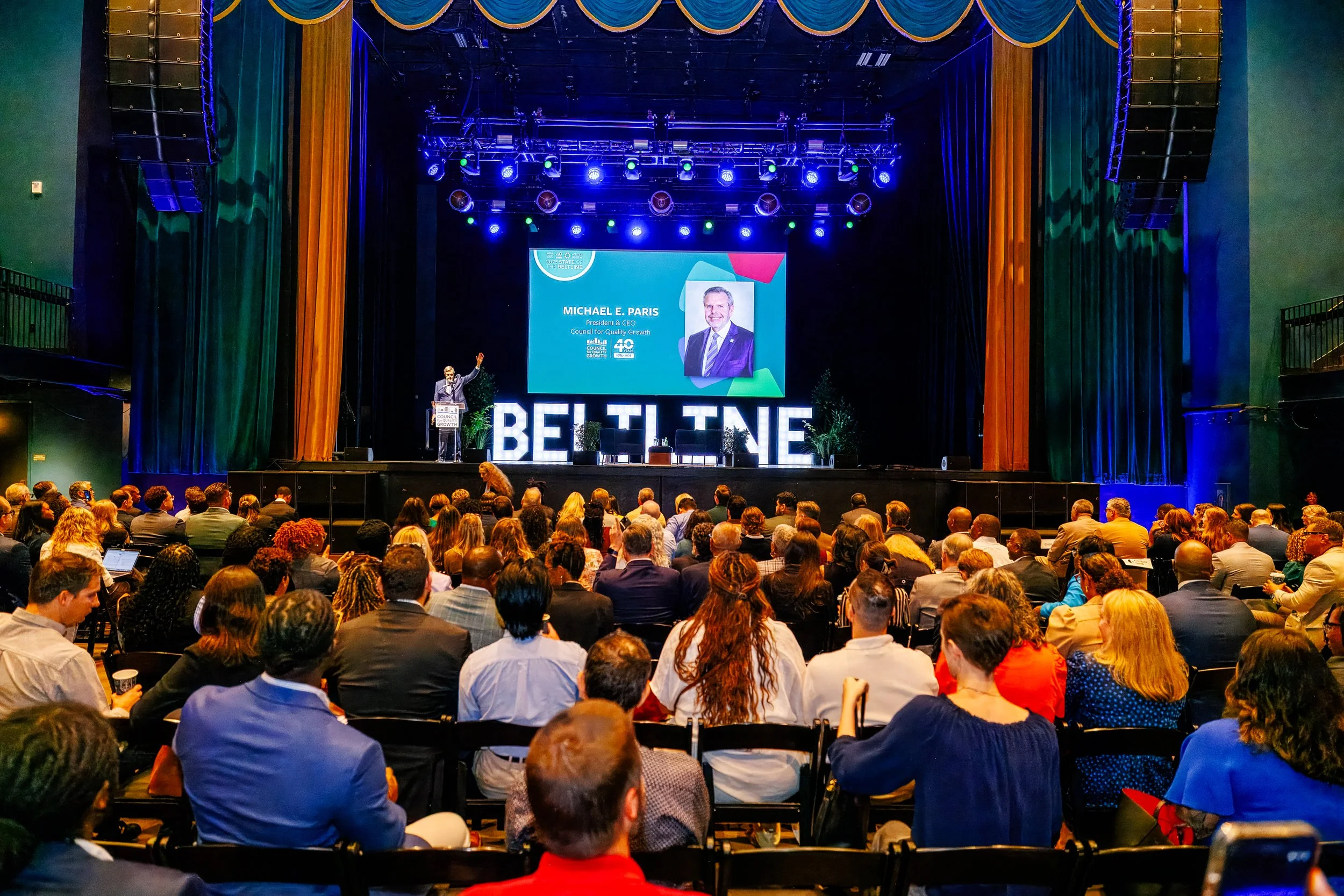 A large indoor conference with a stage and a speaker at the podium. The stage features a large screen displaying the name and photo of Michael E. Paris, along with the words "BELIEVE" spelled out with big illuminated letters. The audience consists of