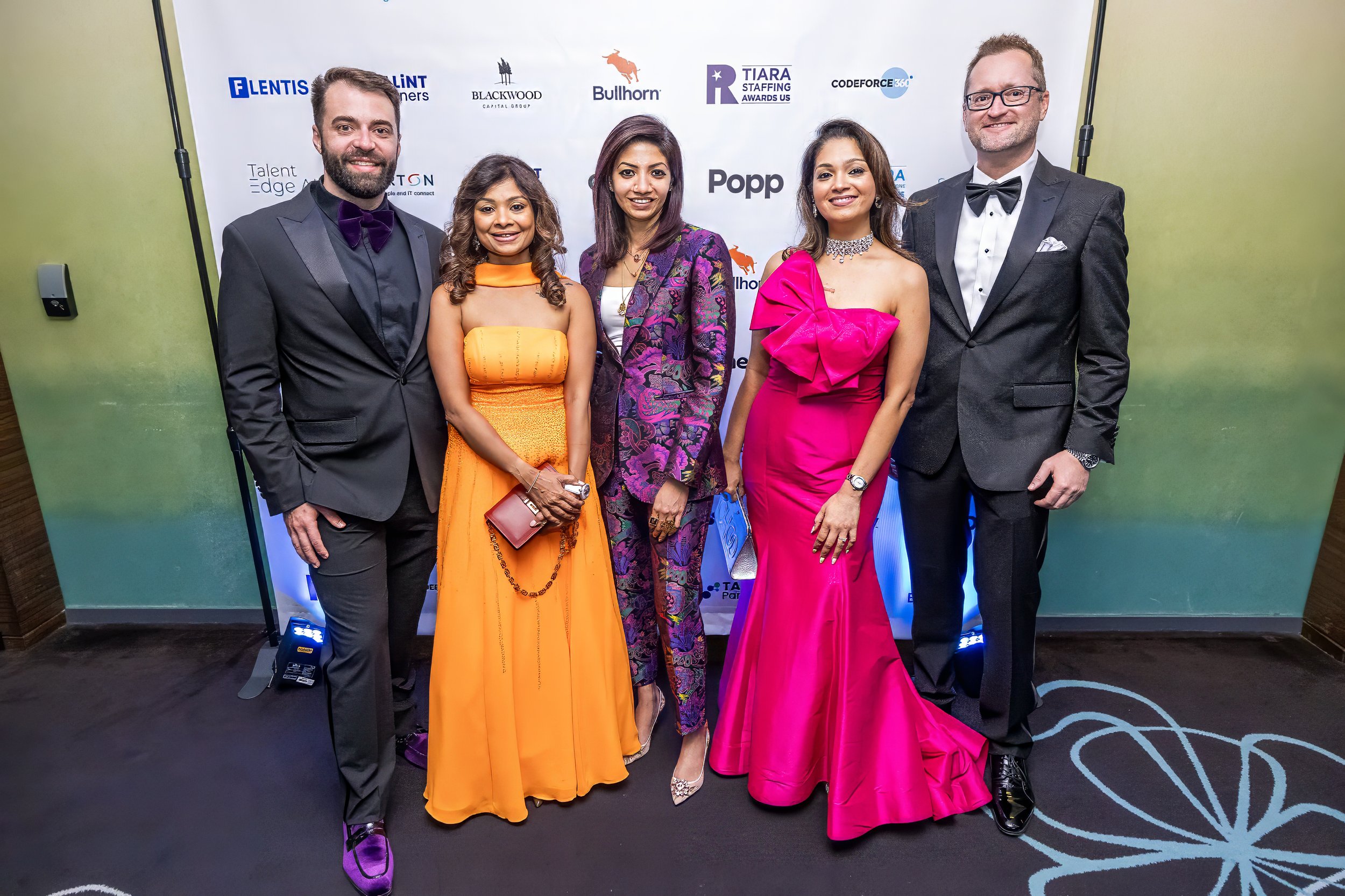 Group of six people dressed in formal attire attending an event, standing in front of a white backdrop with various logos.