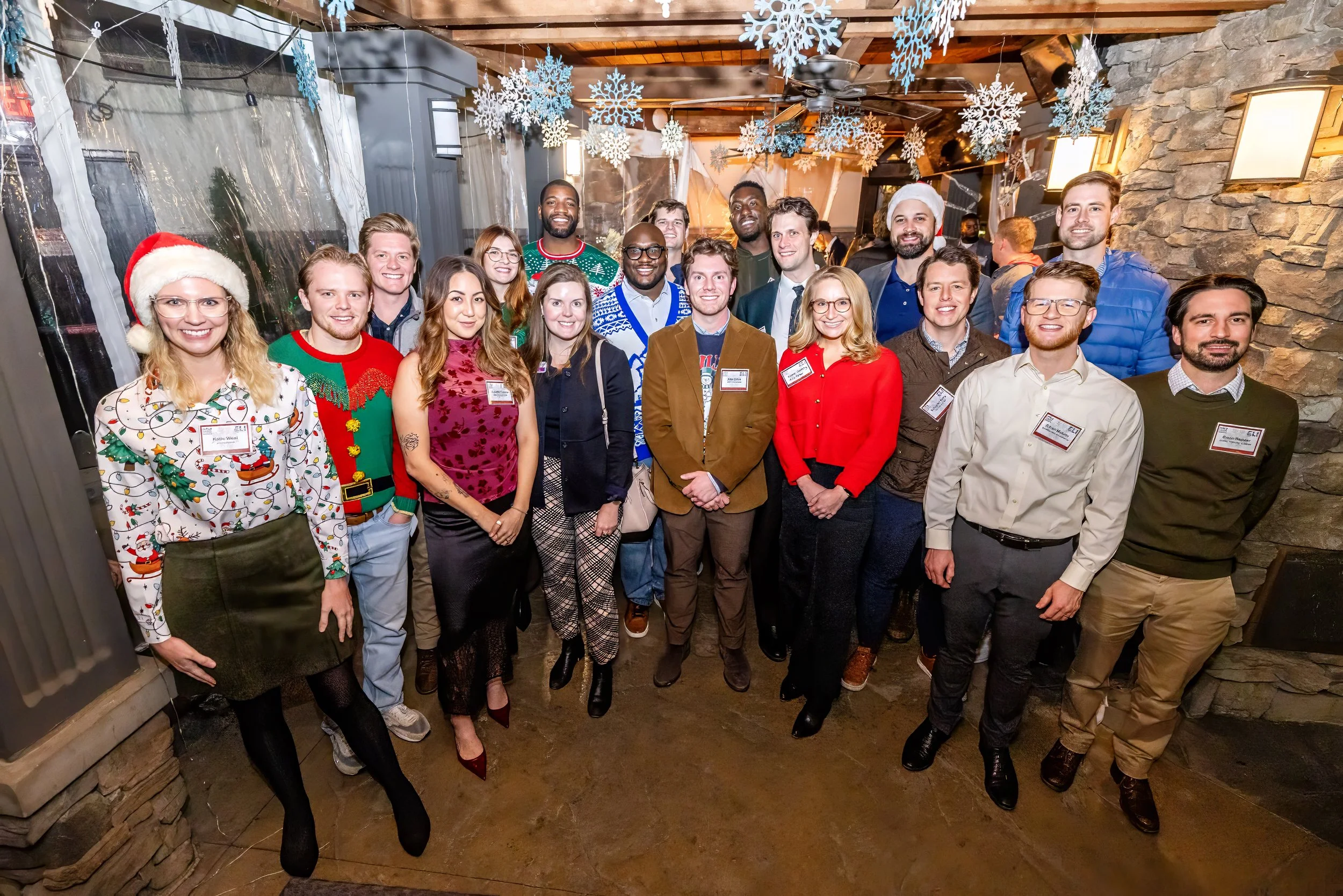 Group of people at a Christmas party, some wearing holiday-themed sweaters and accessories, in a decorated indoor setting with snowflake ornaments hanging from the ceiling.