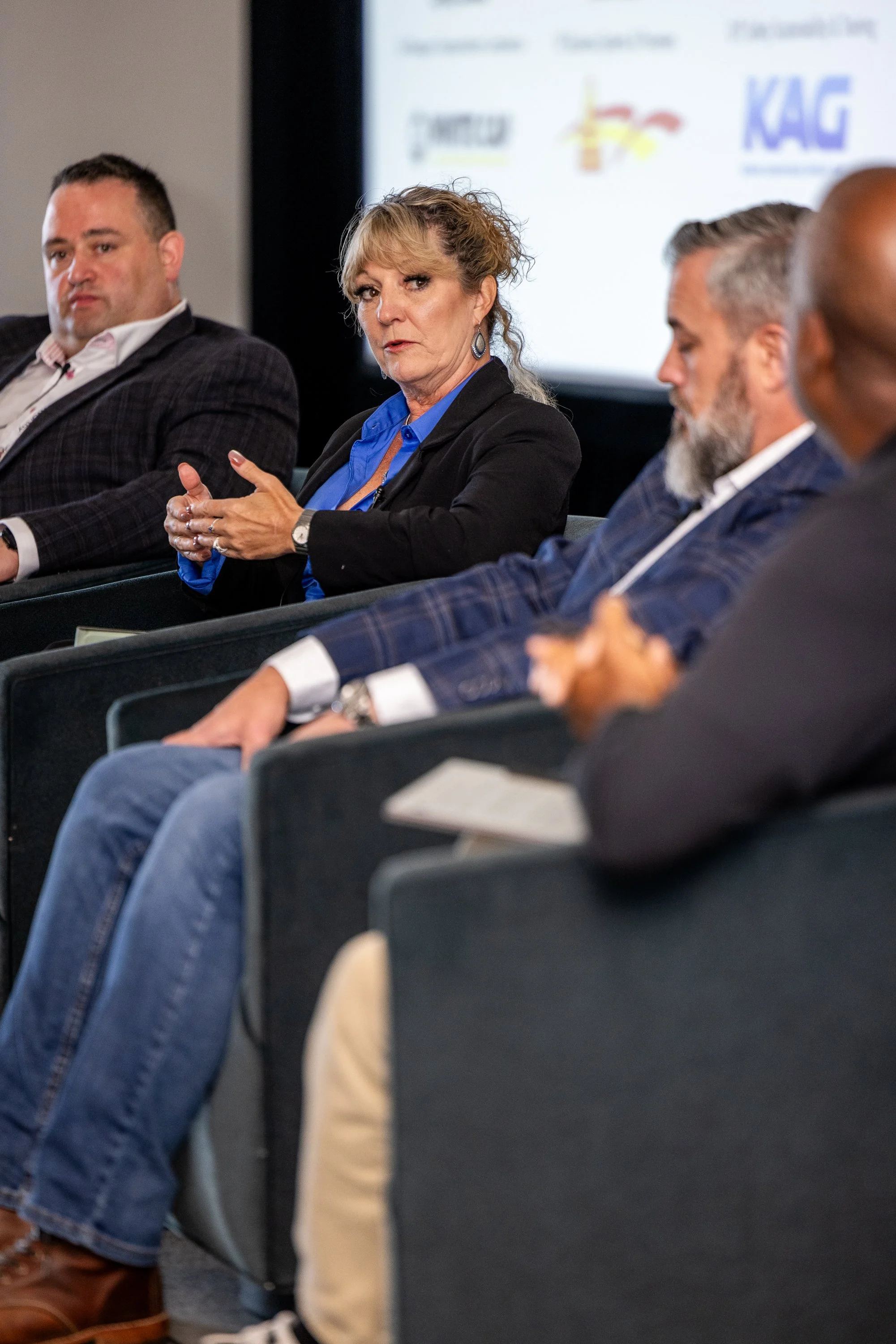 A group of people attending a panel discussion or conference, sitting in chairs and listening. One woman in the center appears to be speaking or making a point. There is a large screen with logos in the background.