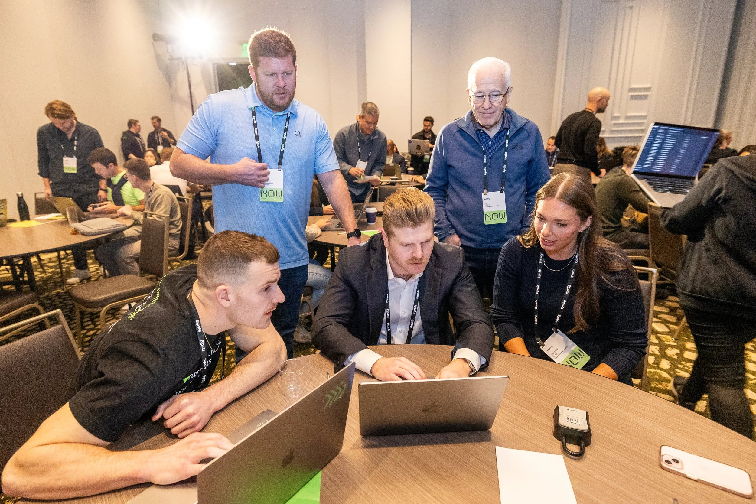 Group of five people gathered around a conference table with laptops, discussing something, at a professional event.
