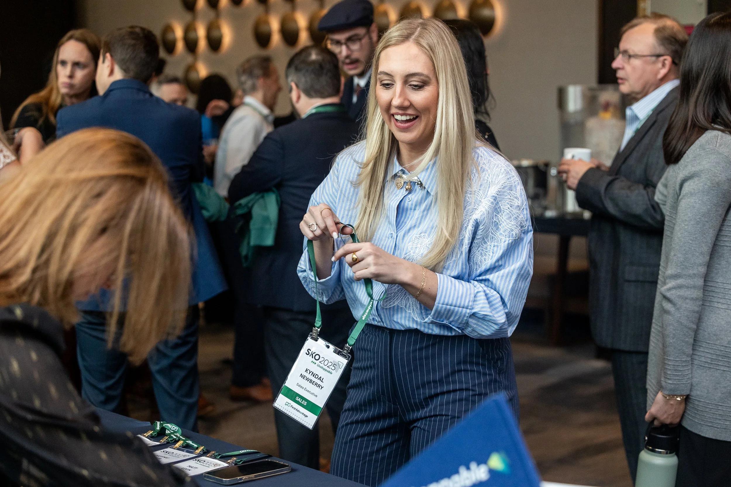 A woman with long blonde hair wearing a light blue shirt and striped pants at a conference, holding a conference badge around her neck, smiling and talking to someone across a table. The background shows other people mingling.