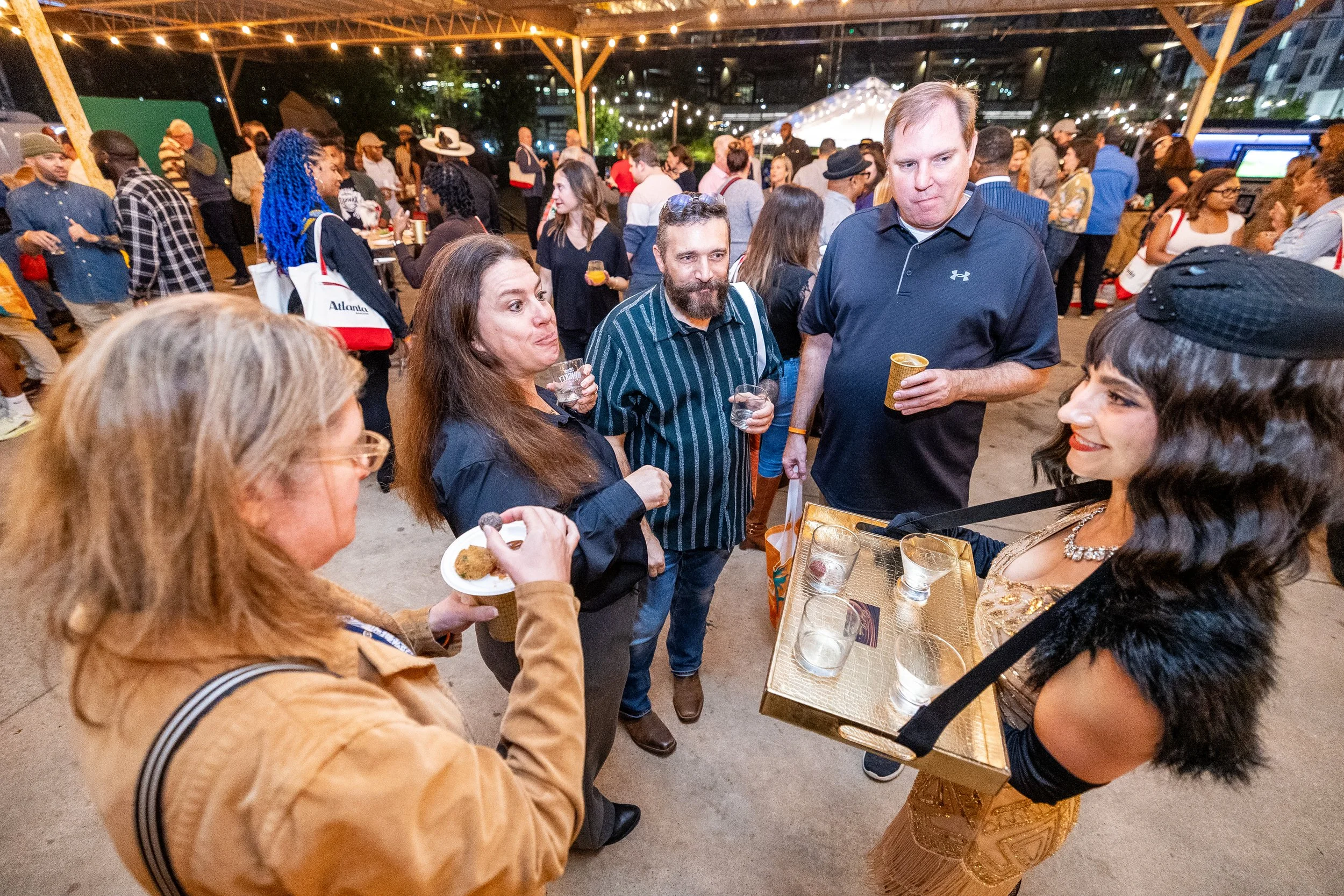People socializing at an outdoor evening event, with a woman in a gold dress serving drinks to others. Background features string lights and a crowd of diverse attendees.