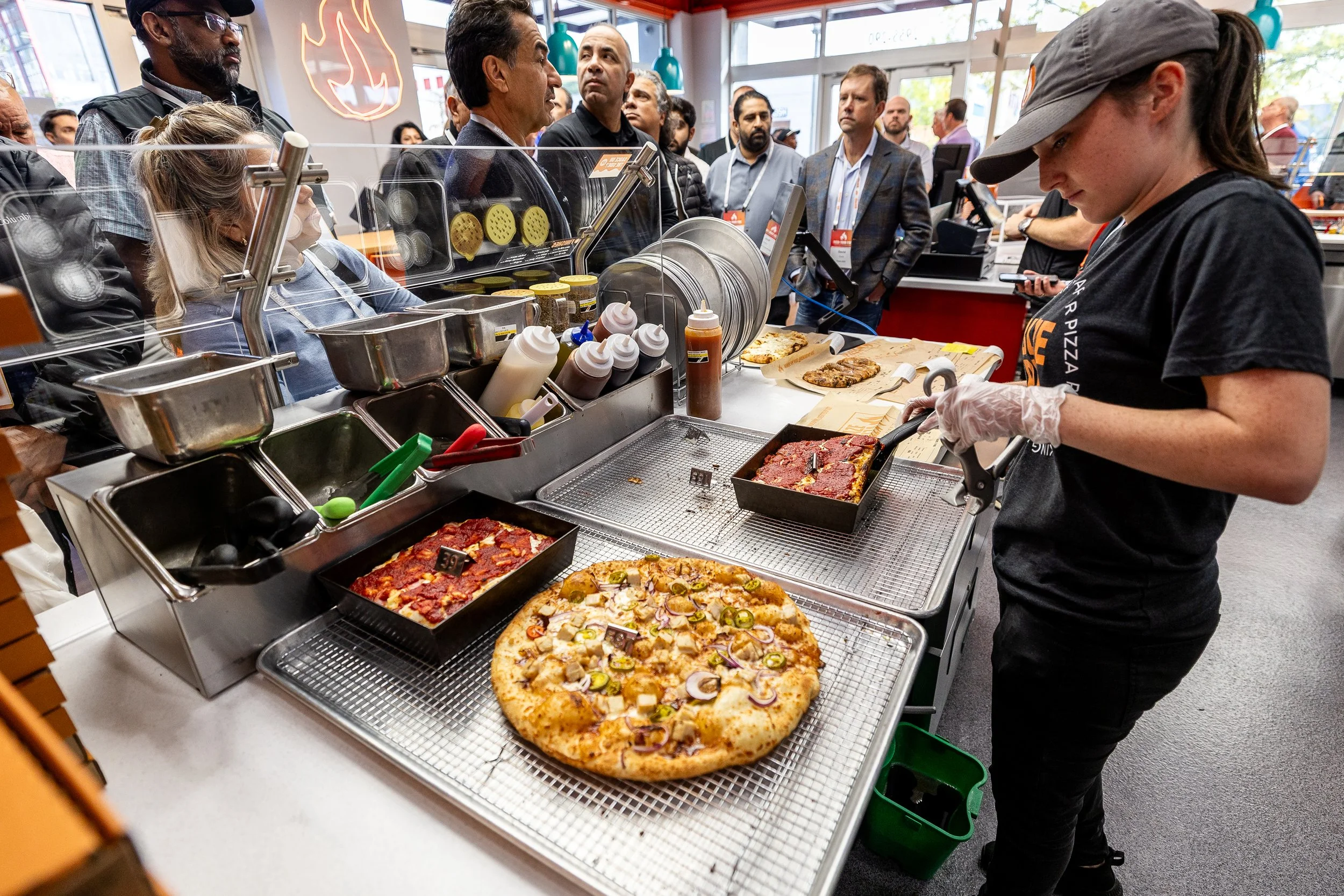 A pizza being prepared at a pizza shop, with various toppings and ingredients visible on the counter. Several people, including a woman in a black shirt and gloves, are working behind the counter, while a group of men waits in front at an event or ma