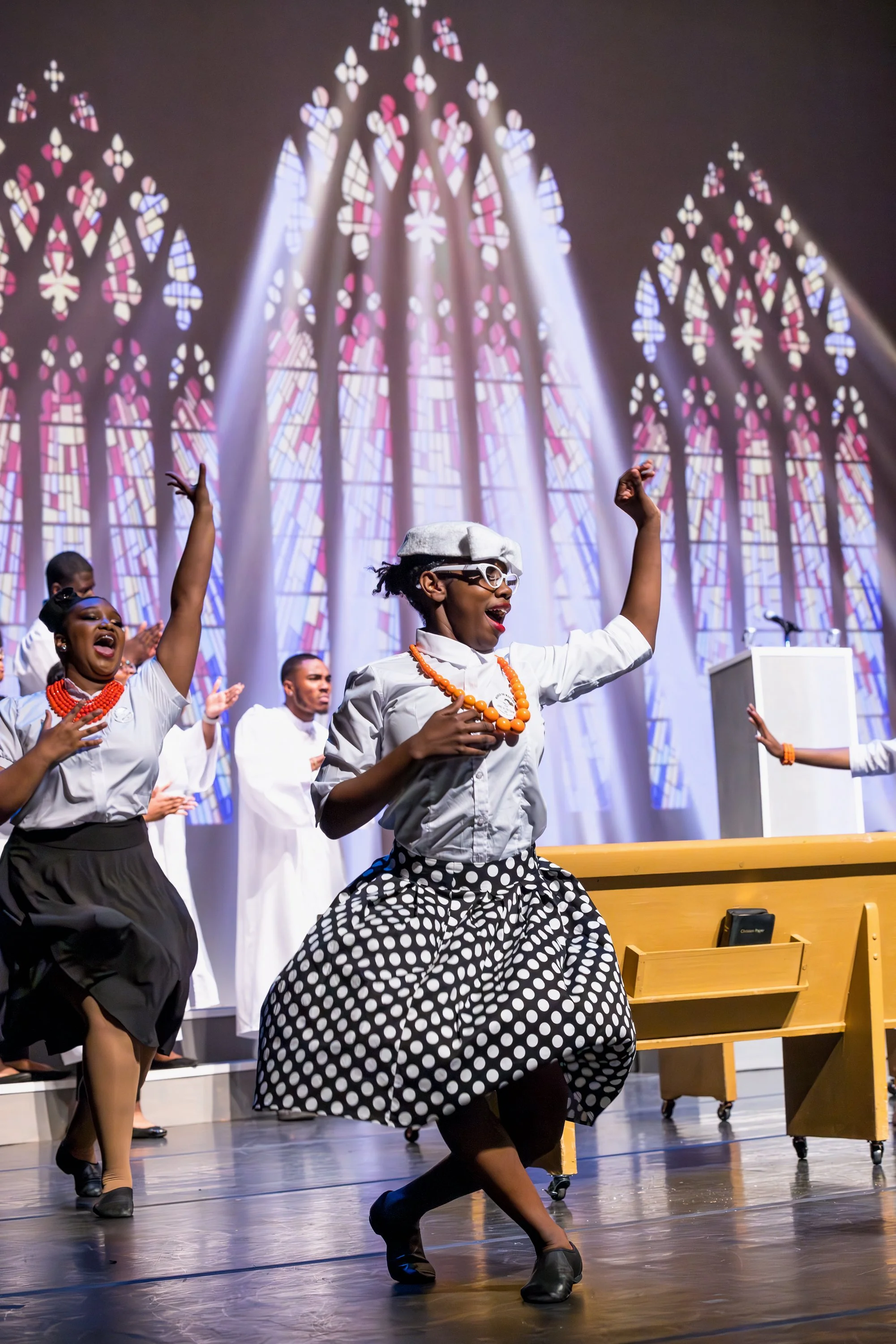 People performing a dance on stage in front of a backdrop of colorful stained glass windows for NCCHR Power to Inspire, some are dressed in white lab coats, and the woman in front wears retro-style glasses, a patterned skirt, and jewelry.