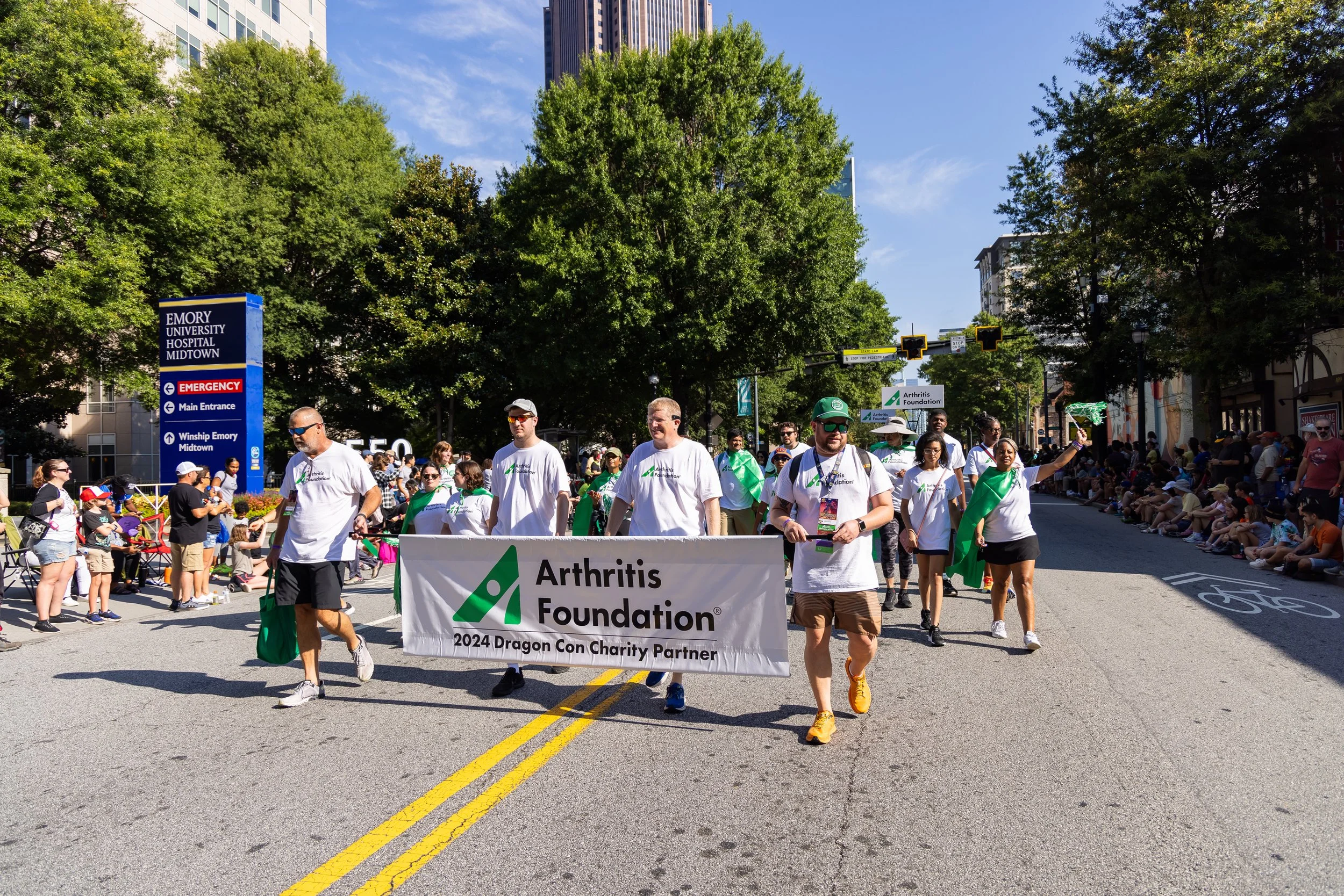 Group of people participating in a charity walk, holding a banner for the Arthritis Foundation during a sunny day in an urban setting with trees and buildings.