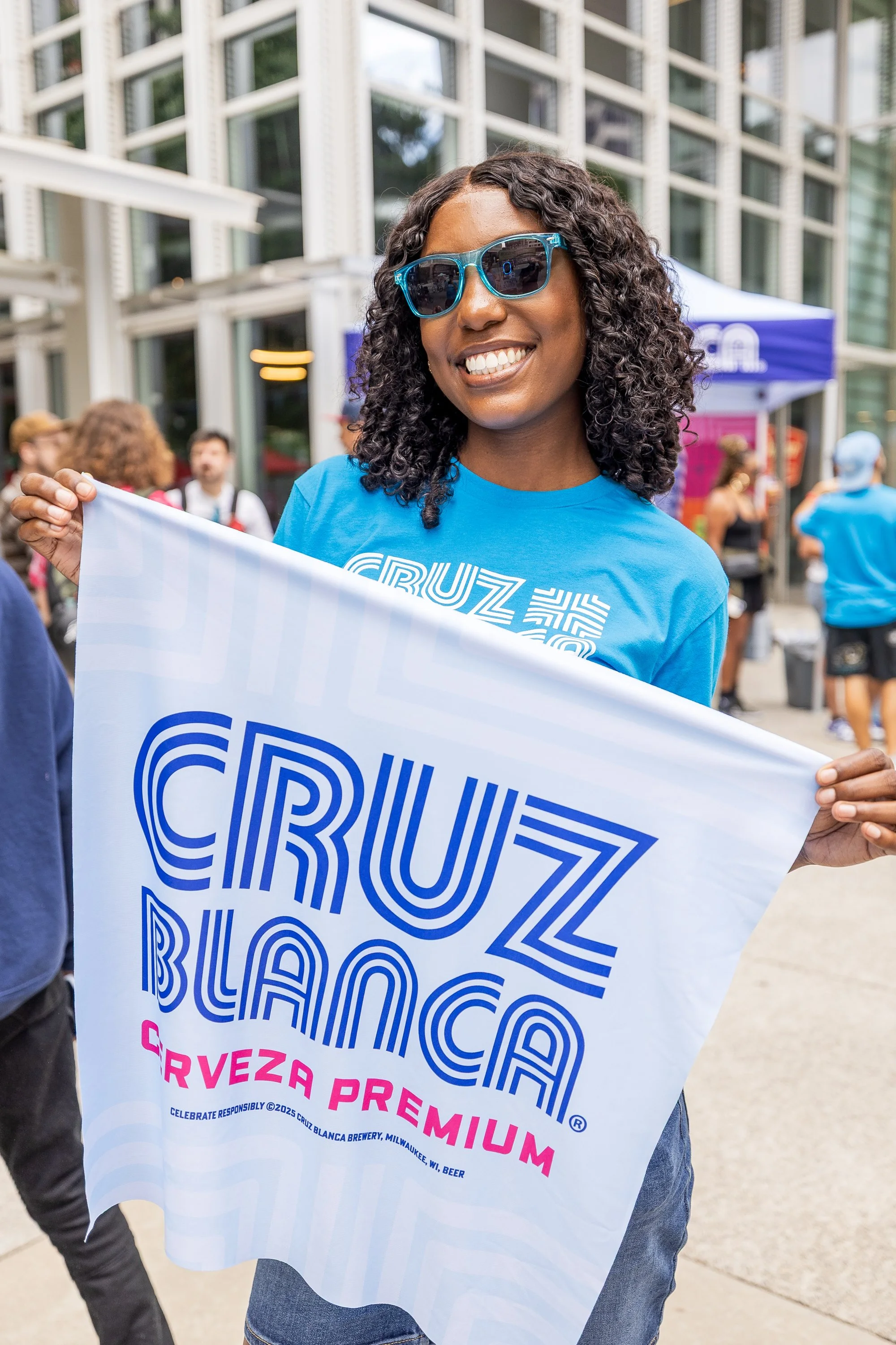 A woman wearing blue sunglasses and a blue Cruz Blanca T-shirt is smiling and holding a Cruz Blanca flag, standing outdoors at a festival or event with people and a tent in the background.