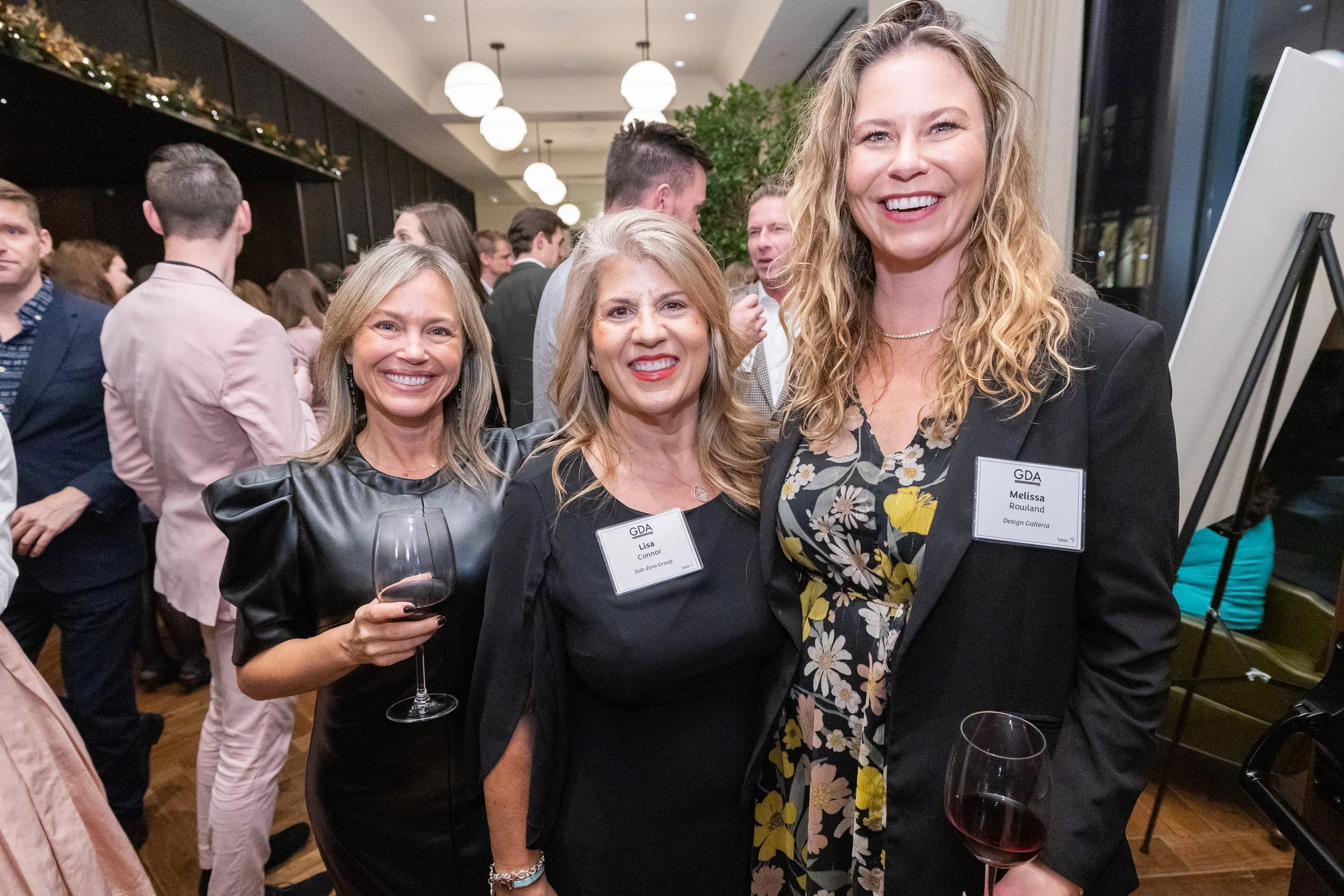 Three women at a social event, smiling and holding wine glasses, wearing formal attire, with a background of other guests and a decorated venue.