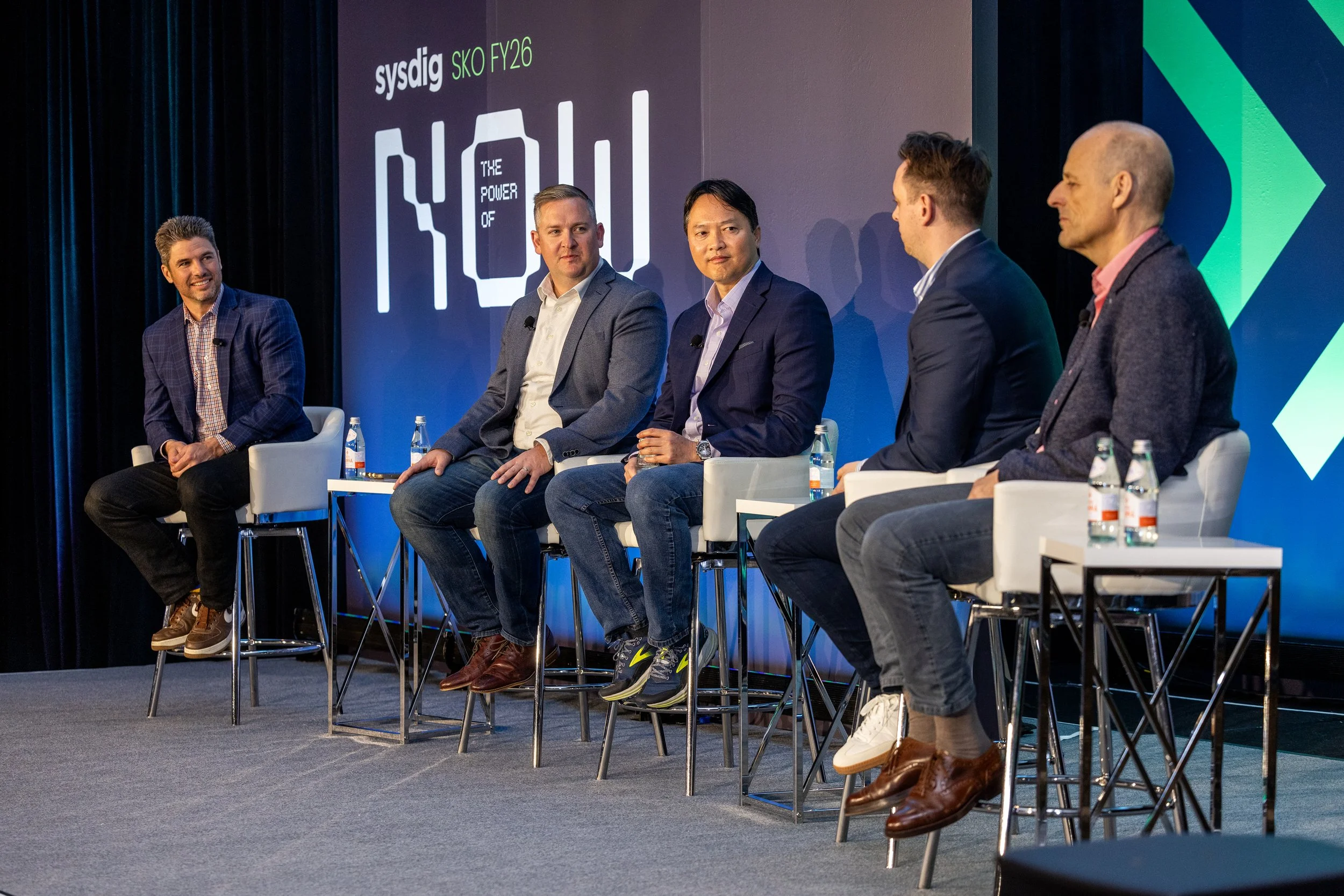 Five men are seated on stage in a panel discussion, with microphones attached to their shirts. The backdrop displays the logo ‘sysdig’ and text ‘SKO FY26’ and ‘The Power of’. Bottled water is placed on small tables beside each person.