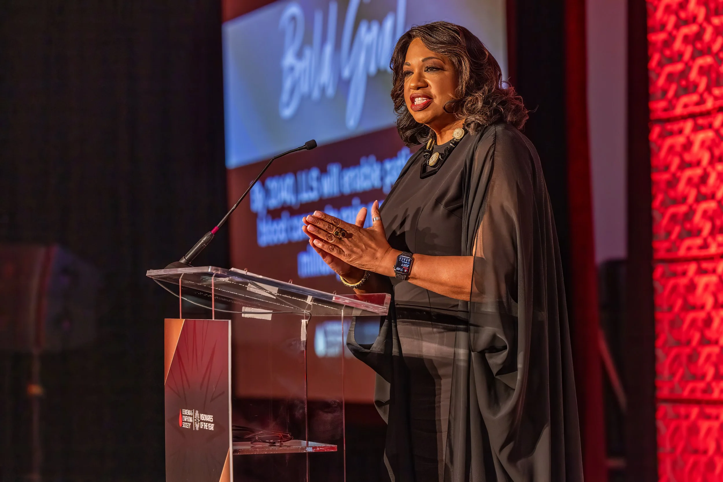 A woman in a black dress giving a speech at a podium on a stage, with a large screen behind her displaying text and a logo.