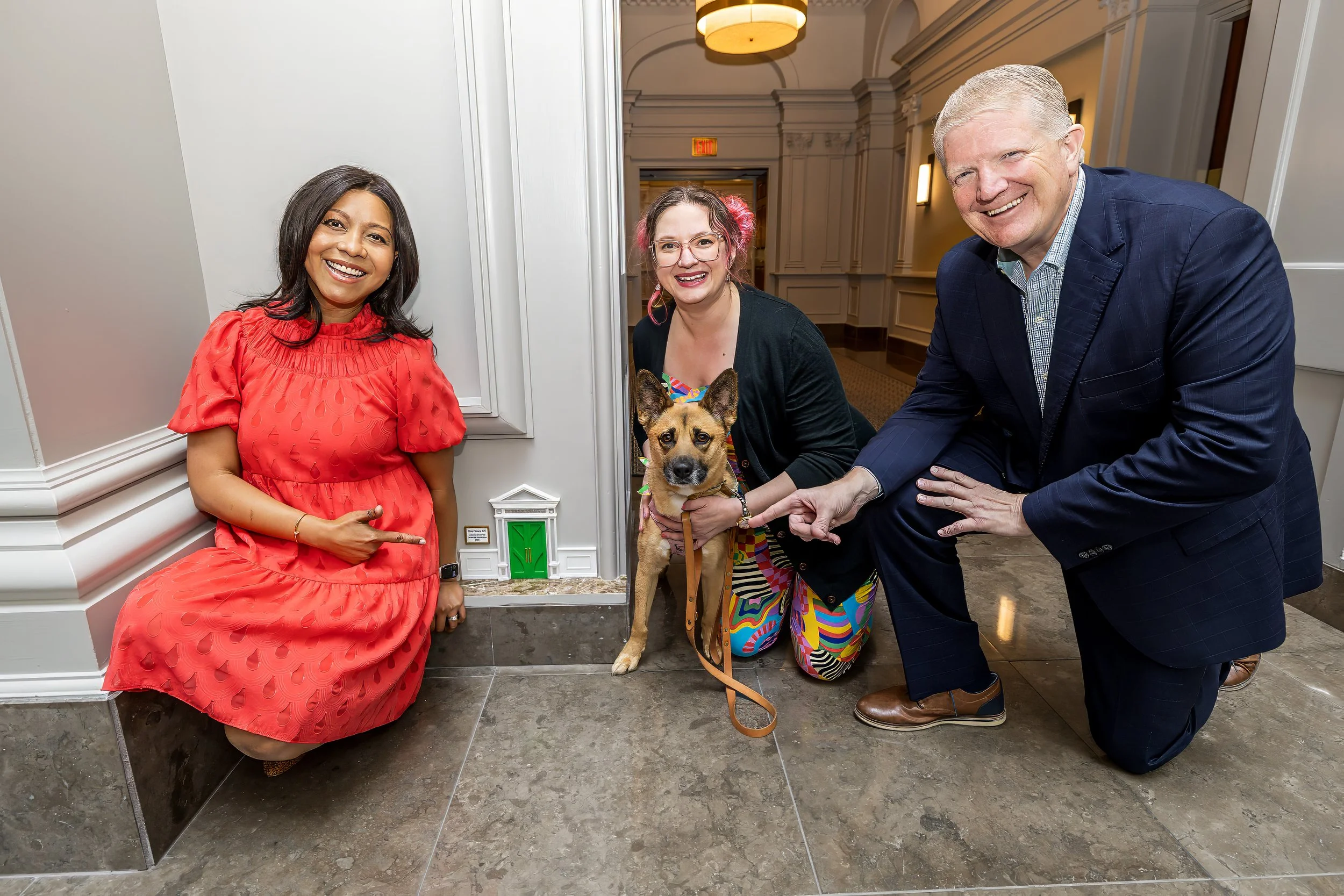 Three people and a dog posing indoors. The woman on the left in a red dress, the woman in the middle with pink hair and colorful skirt holding a dog, and a man in a navy blazer pointing at the dog. The background shows an elegant hallway.