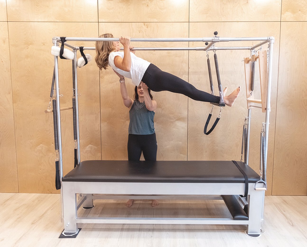 A female client being guided through a Pilates exercise on the Cadillac - a large, versatile piece of Pilates equipment resembling a four-poster bed with a raised mat, surrounded by a metal frame with springs, bars, straps, and other attachments.