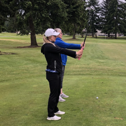 Two women practicing golf on a golf course with trees in the background, one holding a golf club and the other observing.