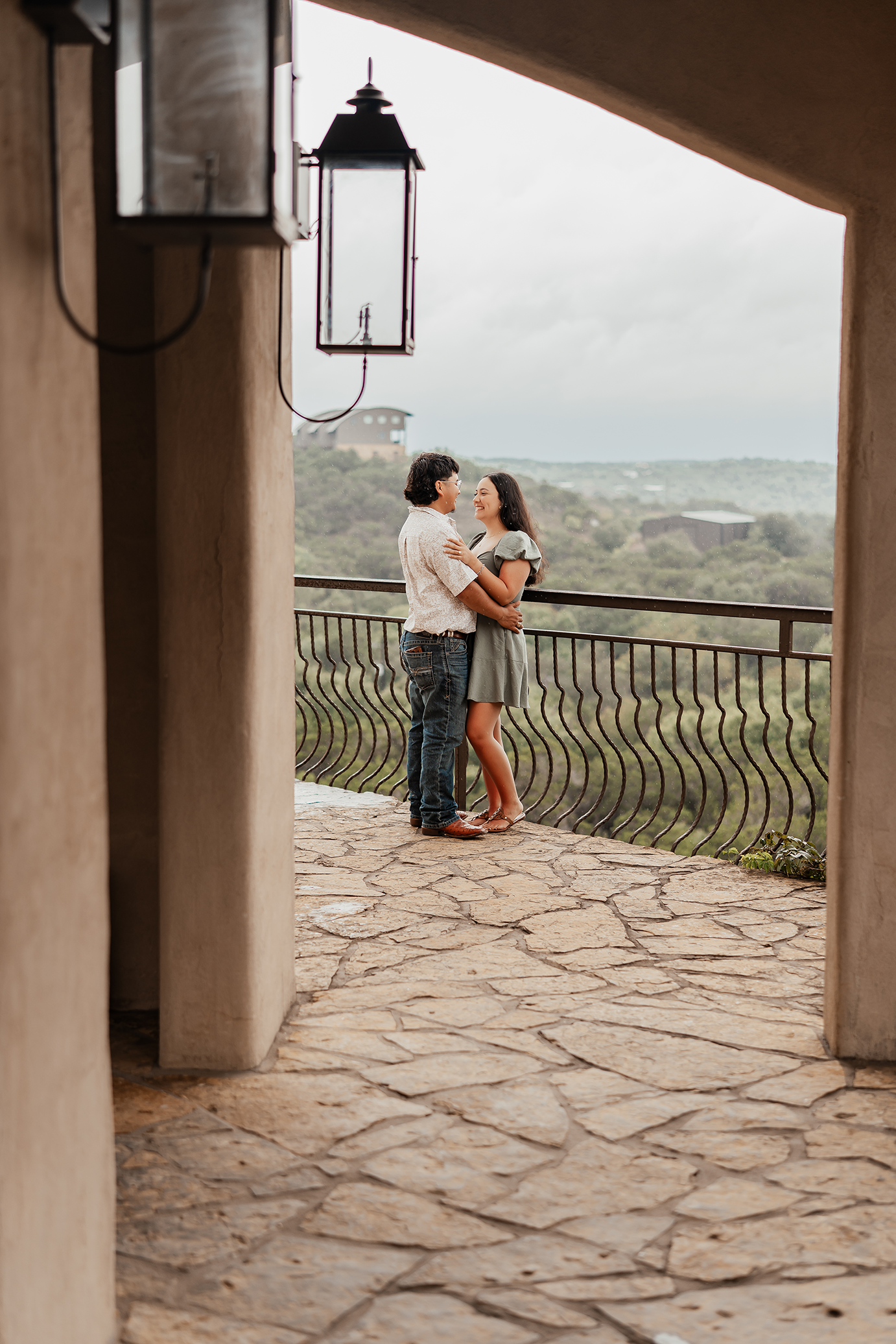 A couple stands close to each other on a balcony overlooking a scenic landscape with trees and buildings, smiling and holding each other.