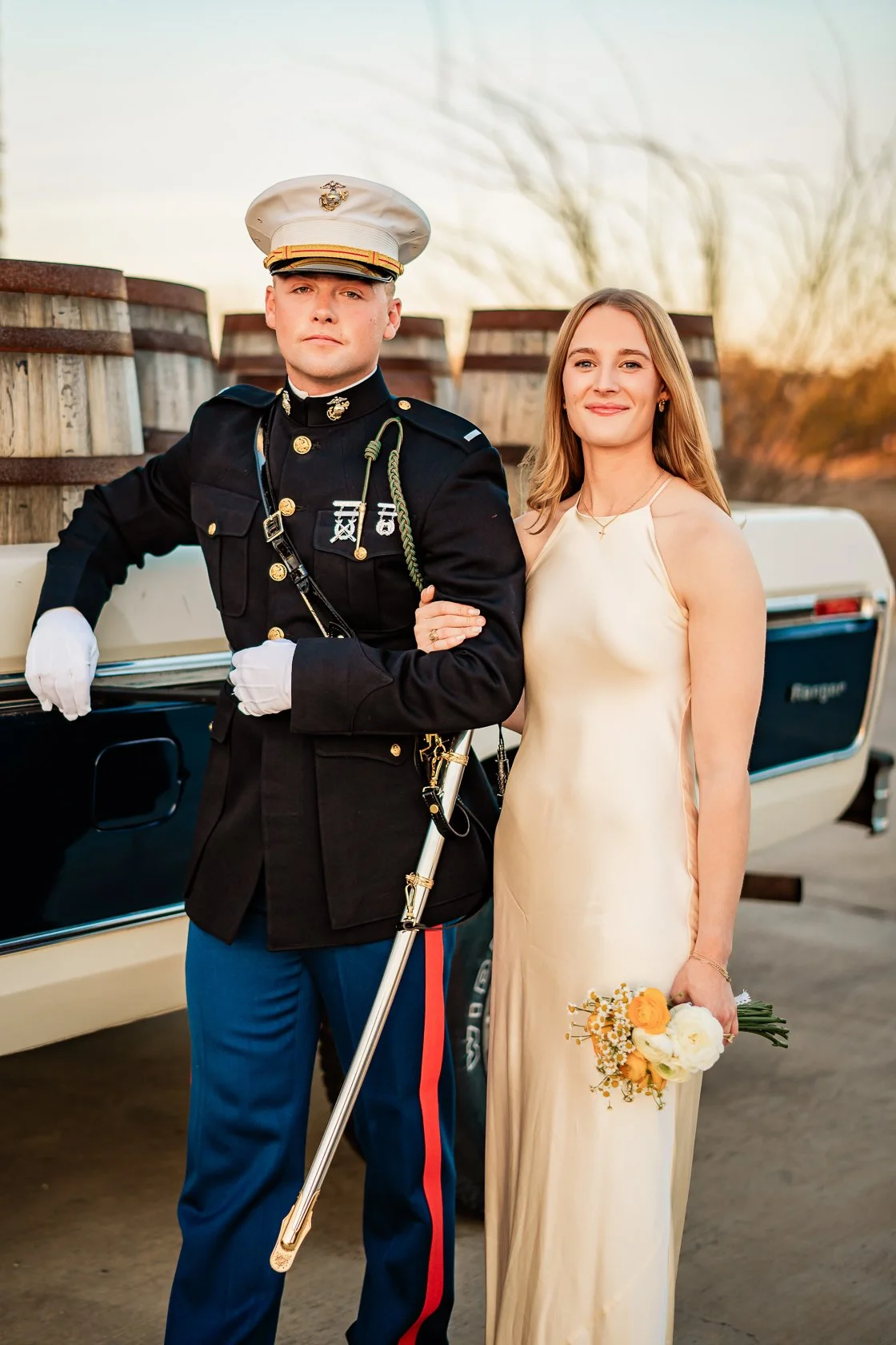 A man in a military uniform and a woman in a cream-colored dress standing outdoors, with a white car and cardboard cartons in the background. The woman is holding a bouquet of flowers.
