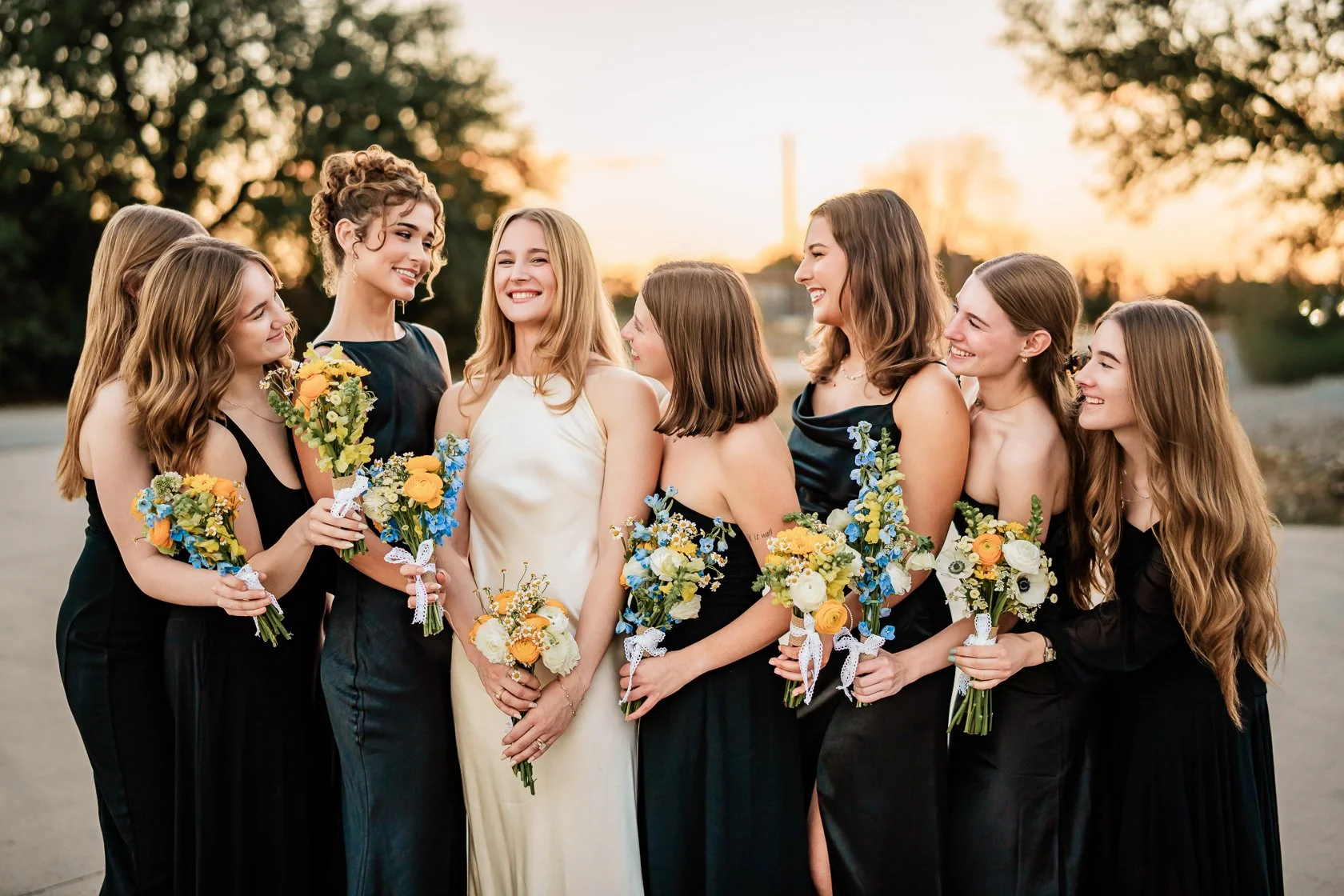 A bride with five bridesmaids standing outdoors during sunset, holding bouquets of yellow and white flowers with blue accents, all smiling and looking at each other.