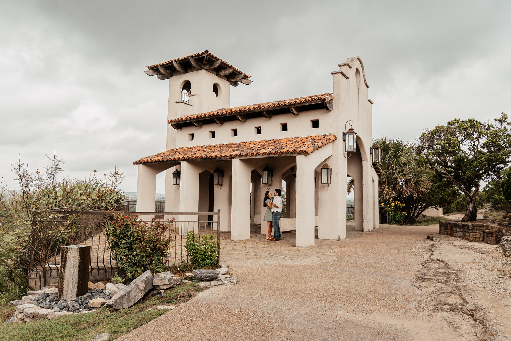 A couple stands under a Spanish-style building with white stucco walls and a red tiled roof, holding each other and smiling outdoors on a cloudy day.