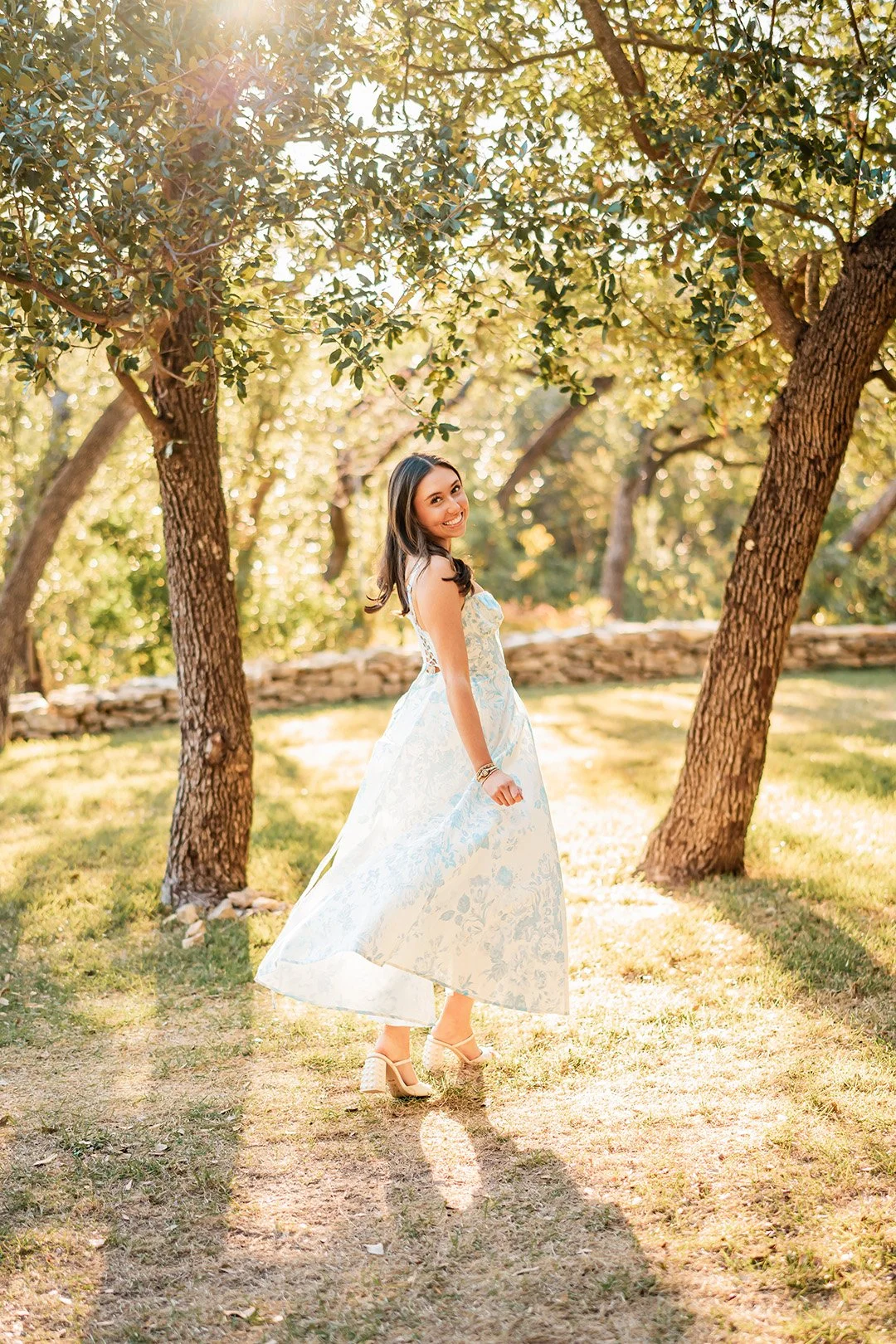 A woman in a white and blue floral dress with high heels, smiling and walking in a sunlit outdoor park with trees and grass.
