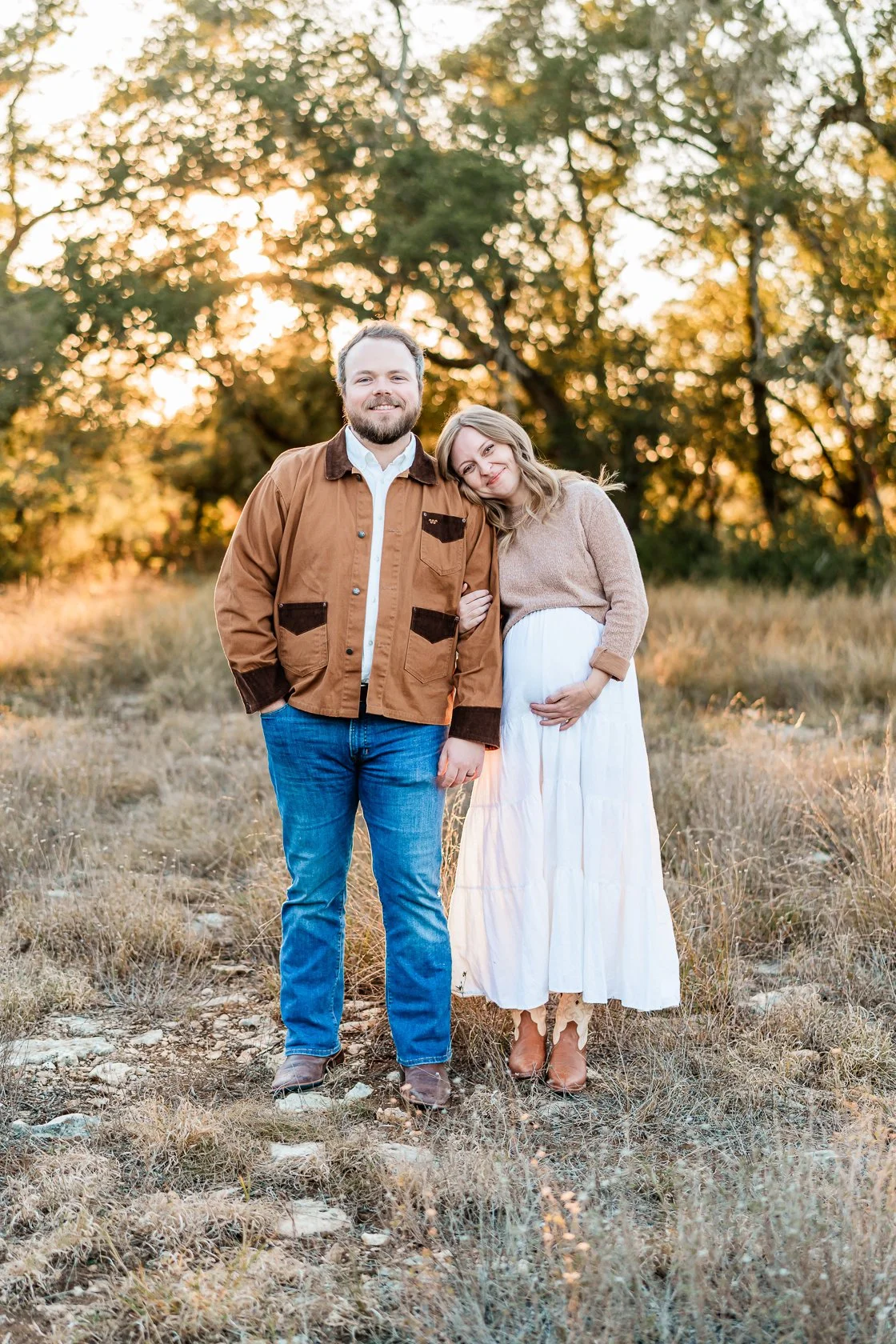 A couple standing outdoors in a field with trees in the background at sunset. The woman is pregnant, wearing a beige sweater and a white skirt, and is resting her head on the man's shoulder. The man is wearing a brown jacket, white shirt, jeans, and 