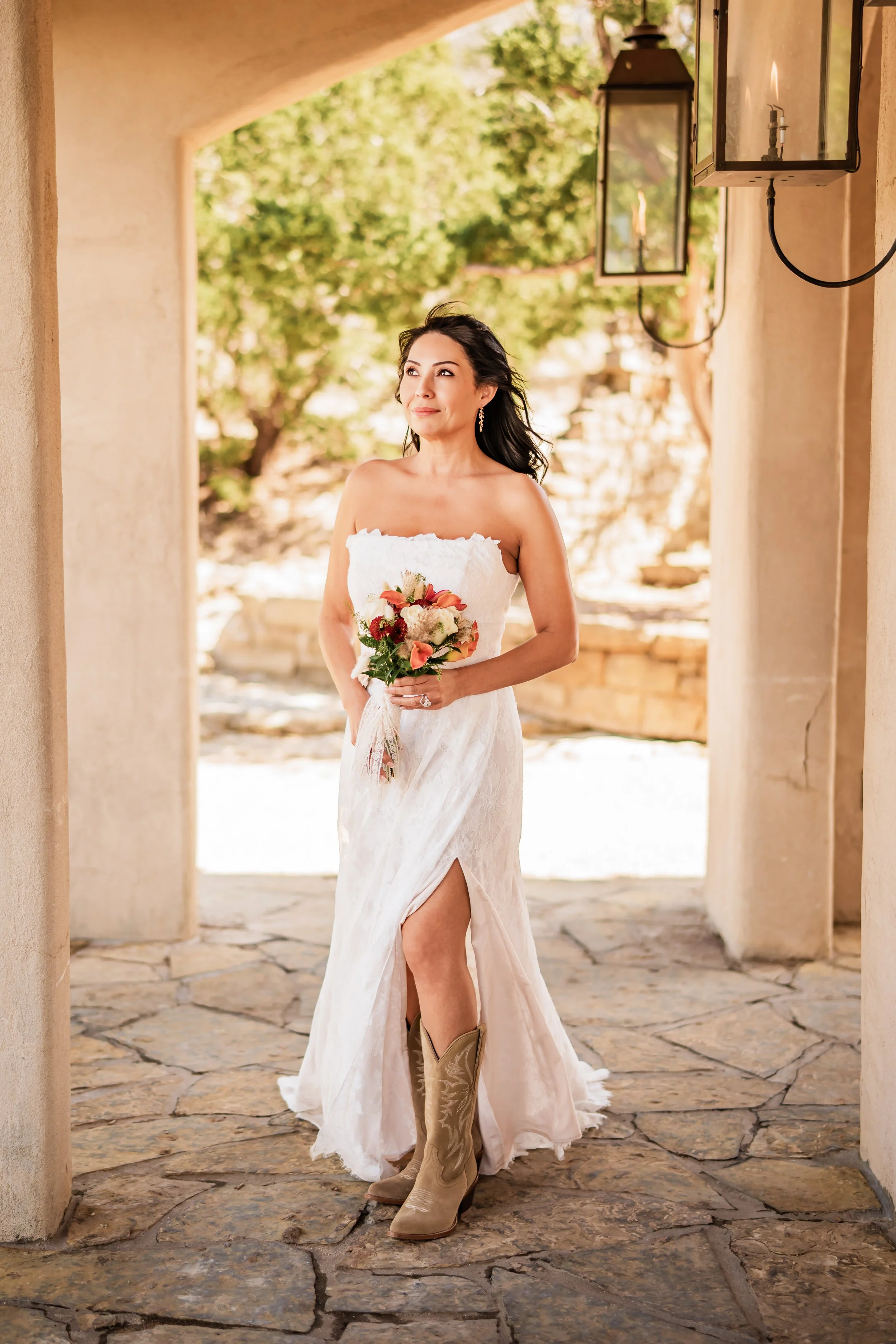 A woman in a strapless white wedding dress with a slit, wearing cowboy boots, holding a bouquet of flowers, standing under an arched stone walkway with hanging lanterns and a background of trees.