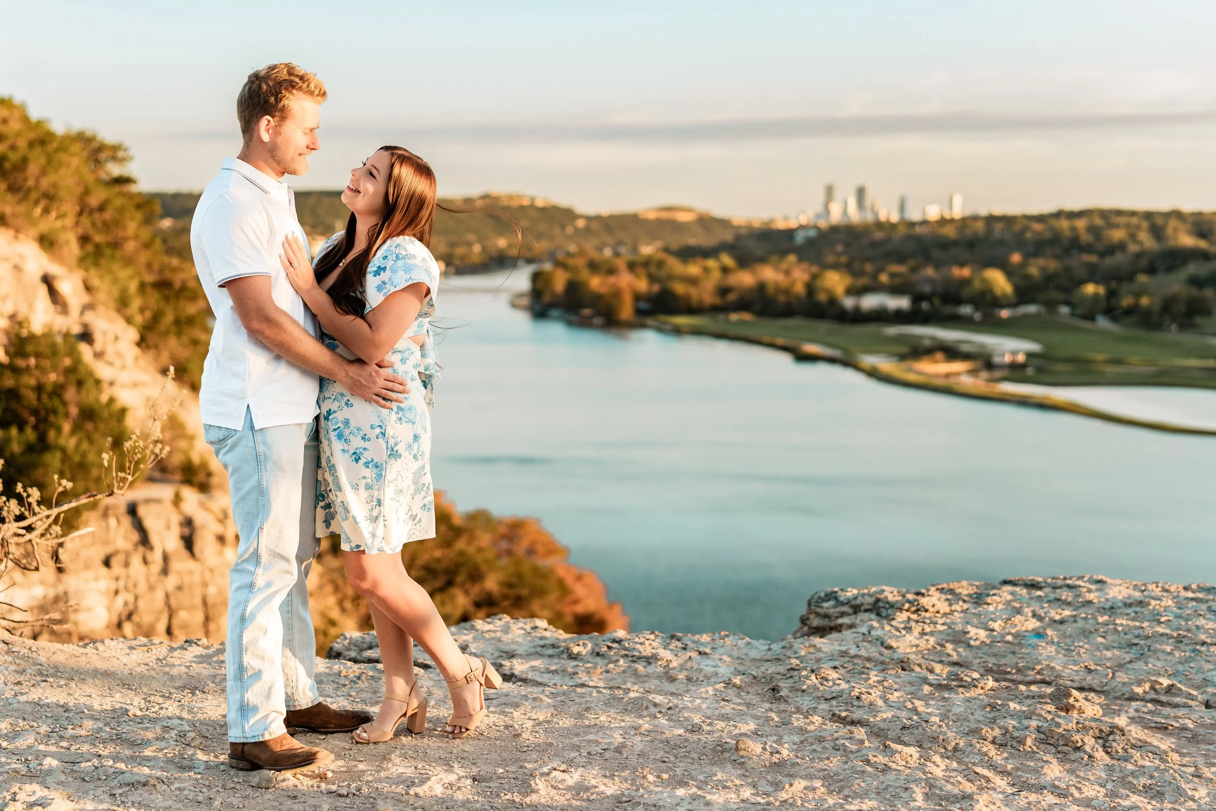 A couple stands close together on rocky ground near a river, smiling and looking into each other's eyes during sunset, with a city skyline visible in the distance.