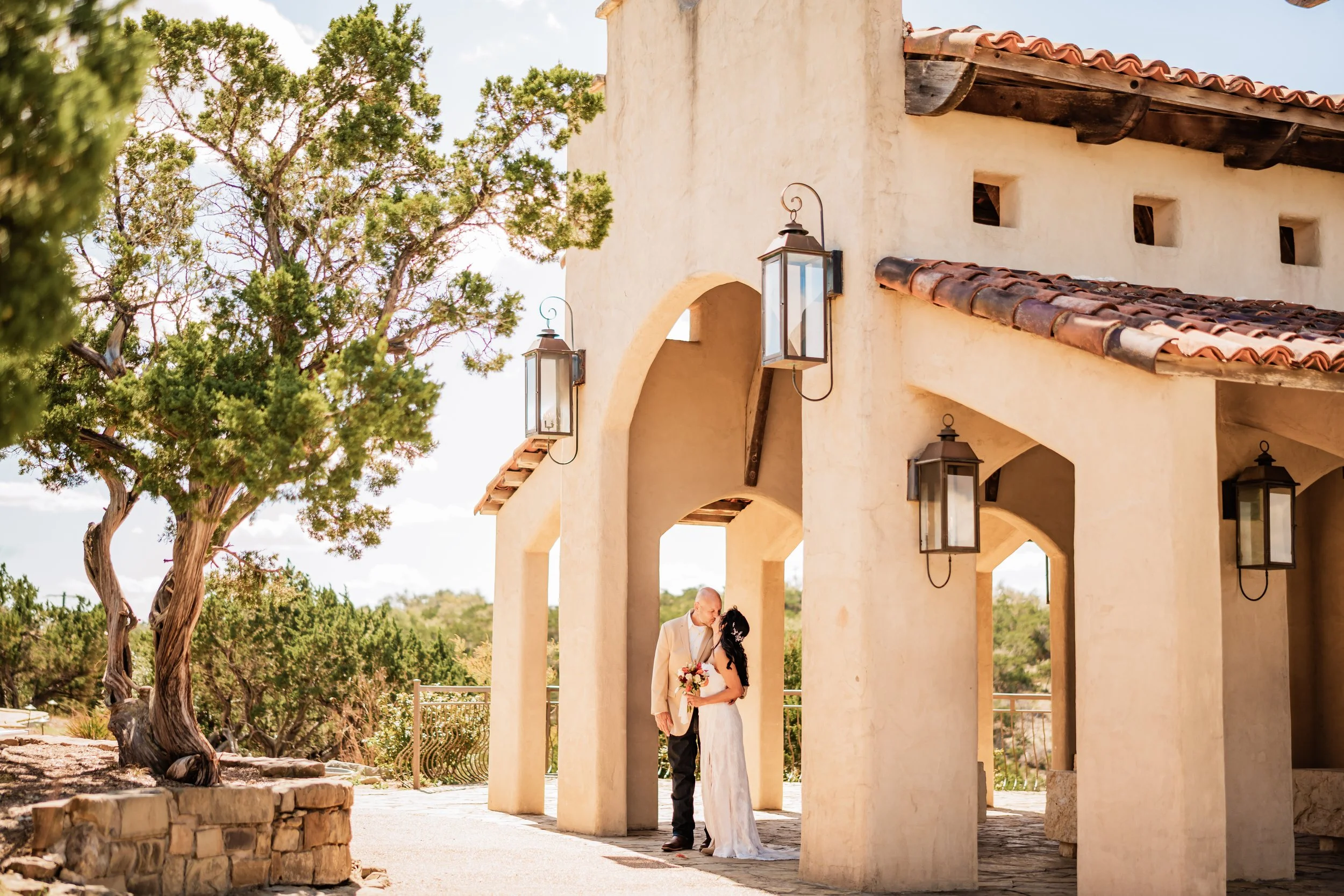 A couple sharing a kiss beneath an arched structure with hanging lanterns in a desert setting with trees.