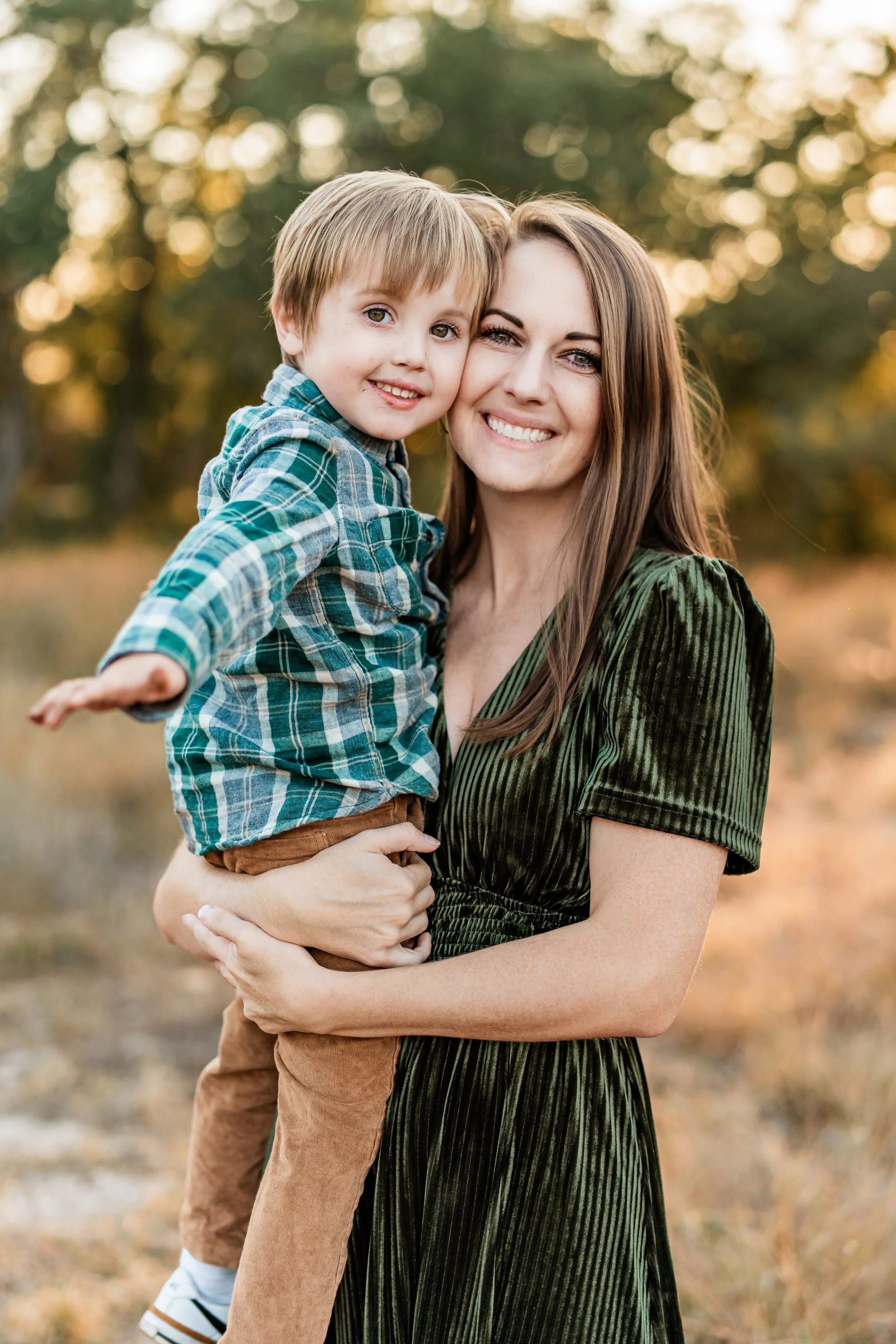 A woman holding a young boy outdoors during golden hour with trees in the background.