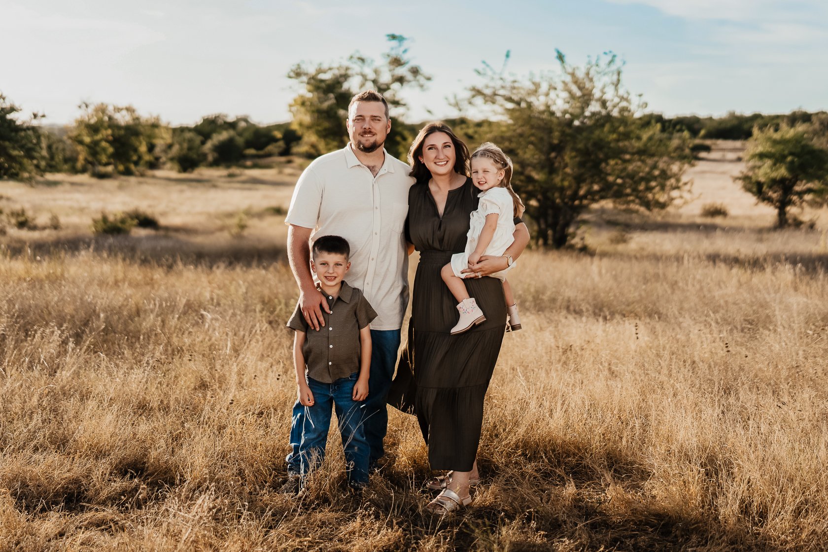 Family of four standing in a grassy field outdoors on a sunny day, with trees in the background, smiling and posing for a photo.