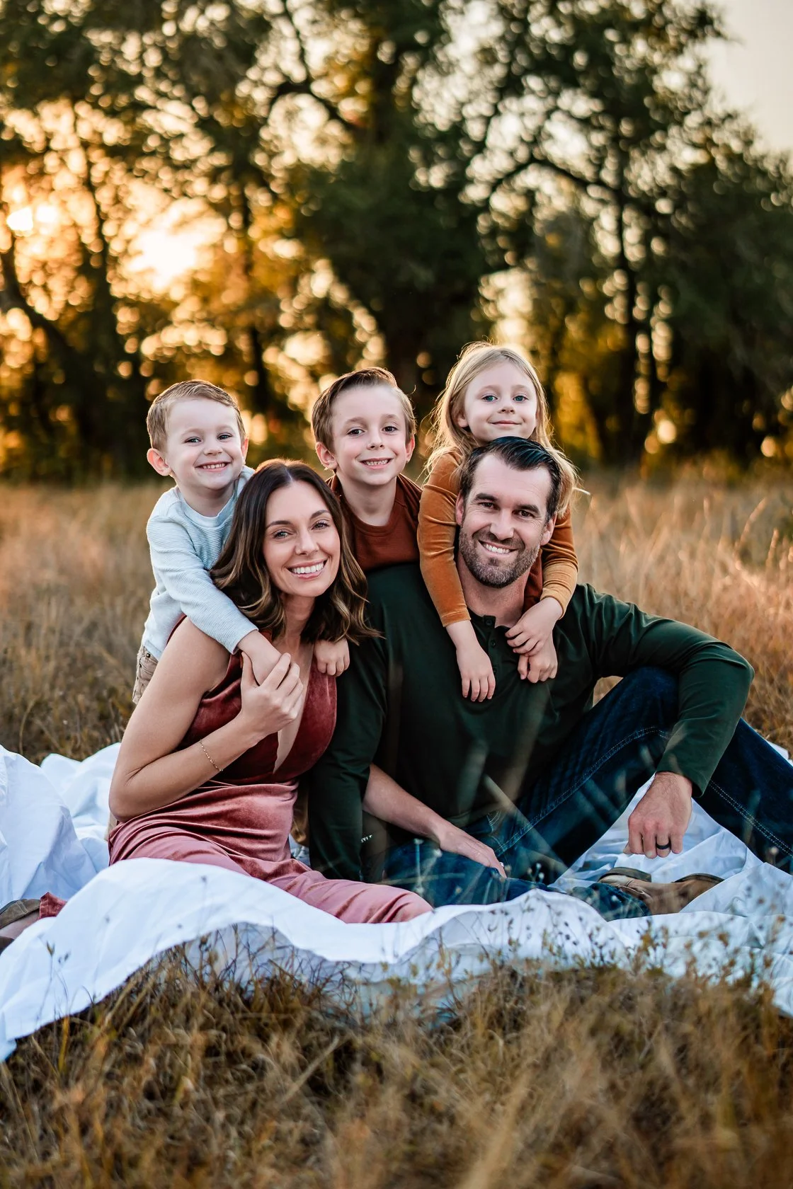 Family of five sitting on a white blanket in a field during sunset, smiling.