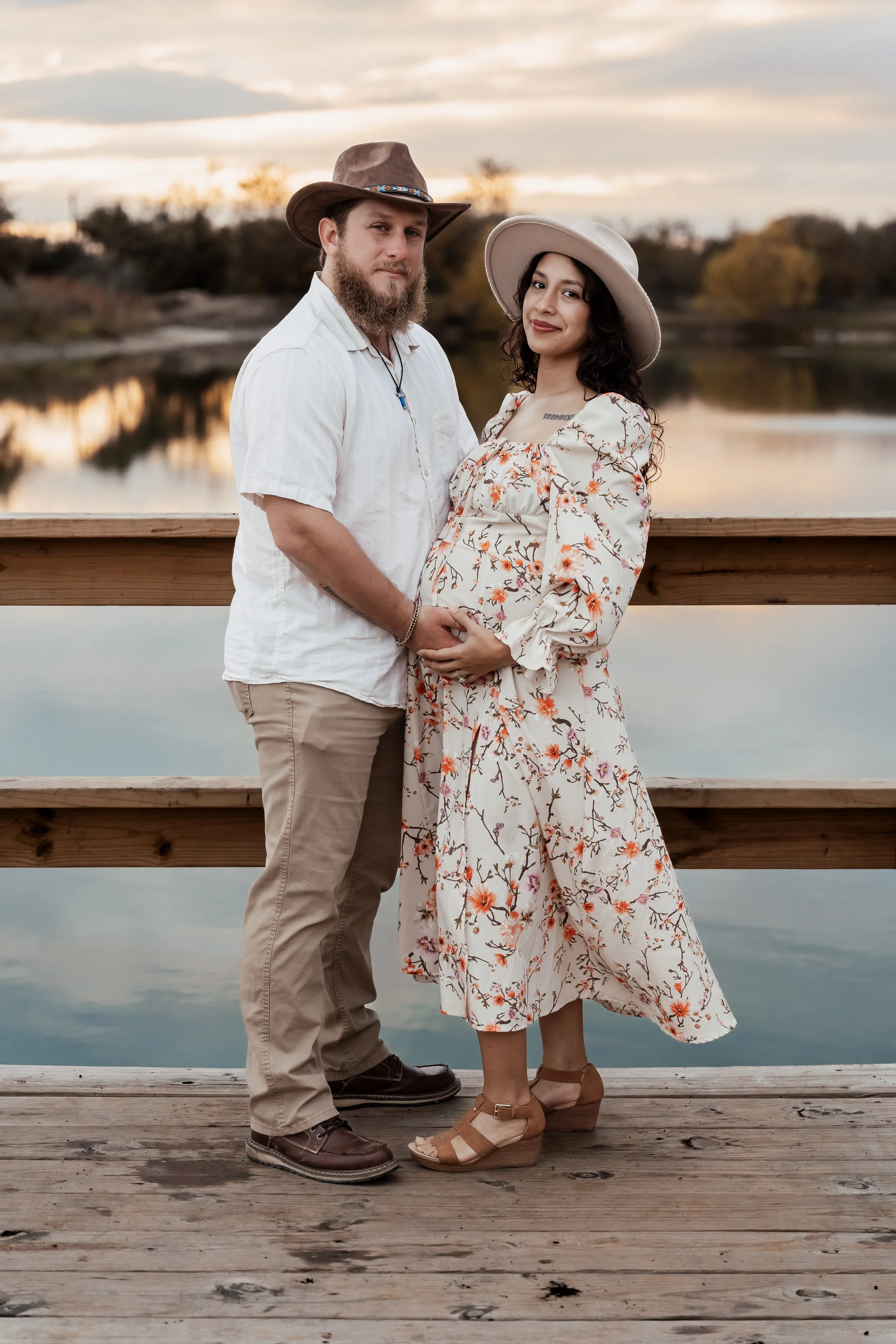 A couple standing on a wooden dock next to a calm lake during sunset. The woman is pregnant and wearing a floral dress and wide-brimmed hat. The man is dressed in a white shirt, beige pants, and a brown hat, holding her belly and gazing at the camera