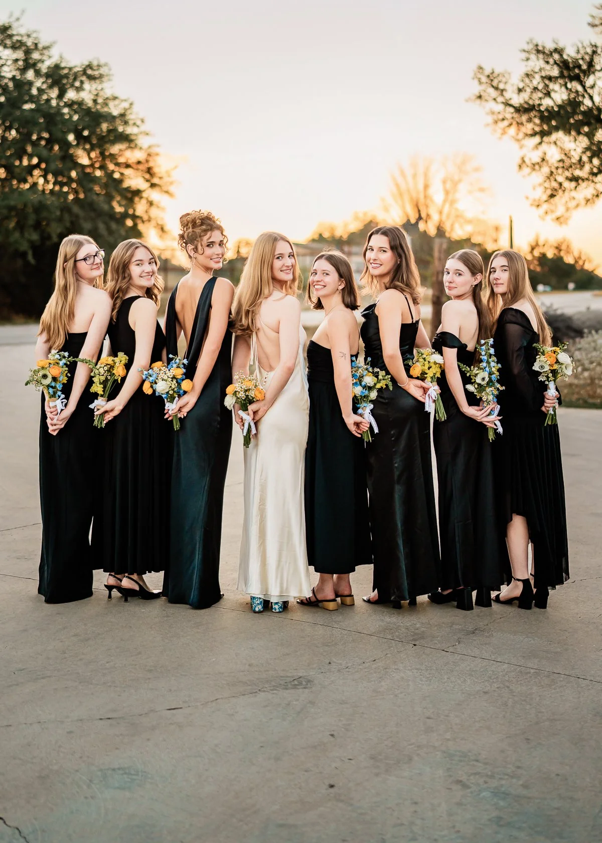 Group of women in formal dresses holding bouquets standing outdoors during sunset.