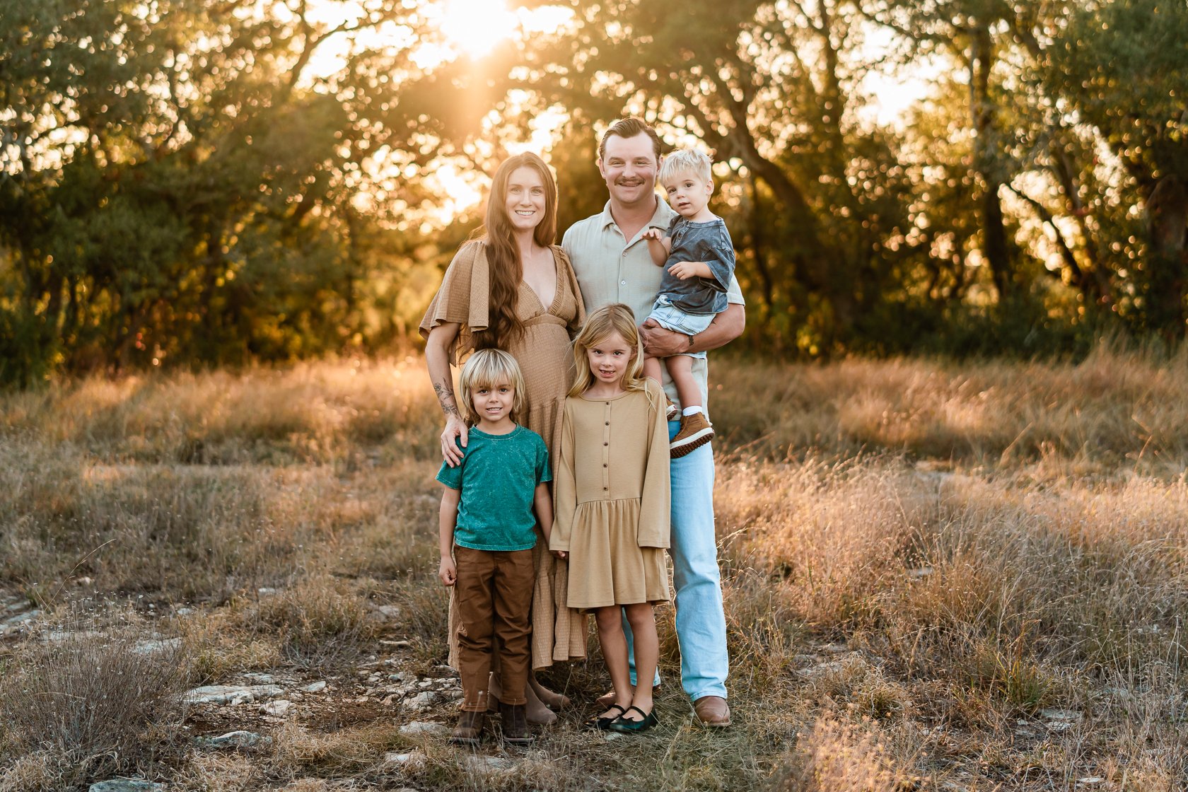 A family of five standing outdoors in a grassy field at sunset, smiling at the camera. The family includes a pregnant woman, a man, and three children, with trees in the background.