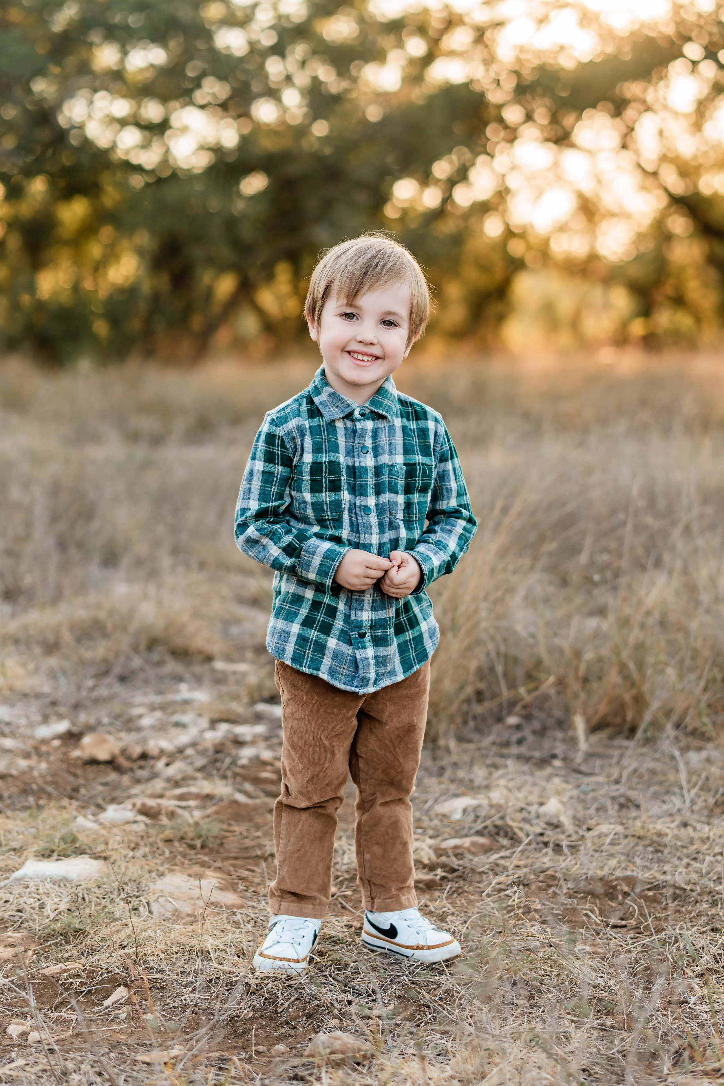 A young boy smiling and standing outdoors on a dirt path in a field with tall grass, with trees in the background and sunlight filtering through the leaves.