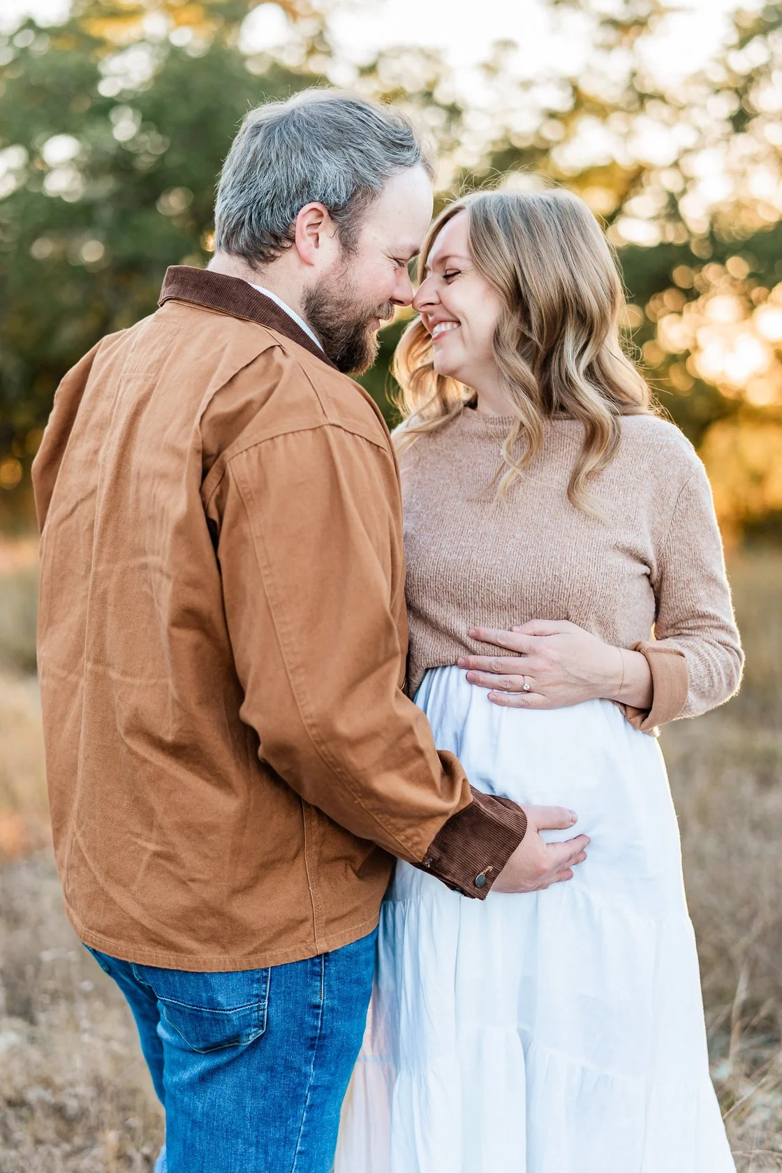 A smiling couple outdoors, facing each other with their foreheads touching, during sunset with trees in the background.