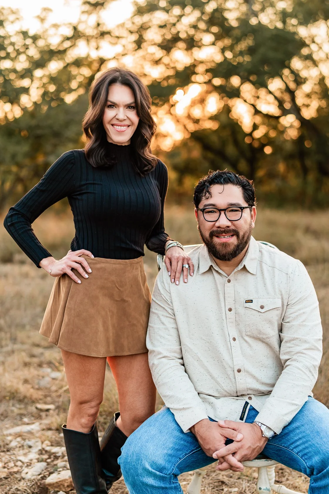 A woman standing with her hand on a man's shoulder, both smiling outdoors during sunset, with trees in the background.