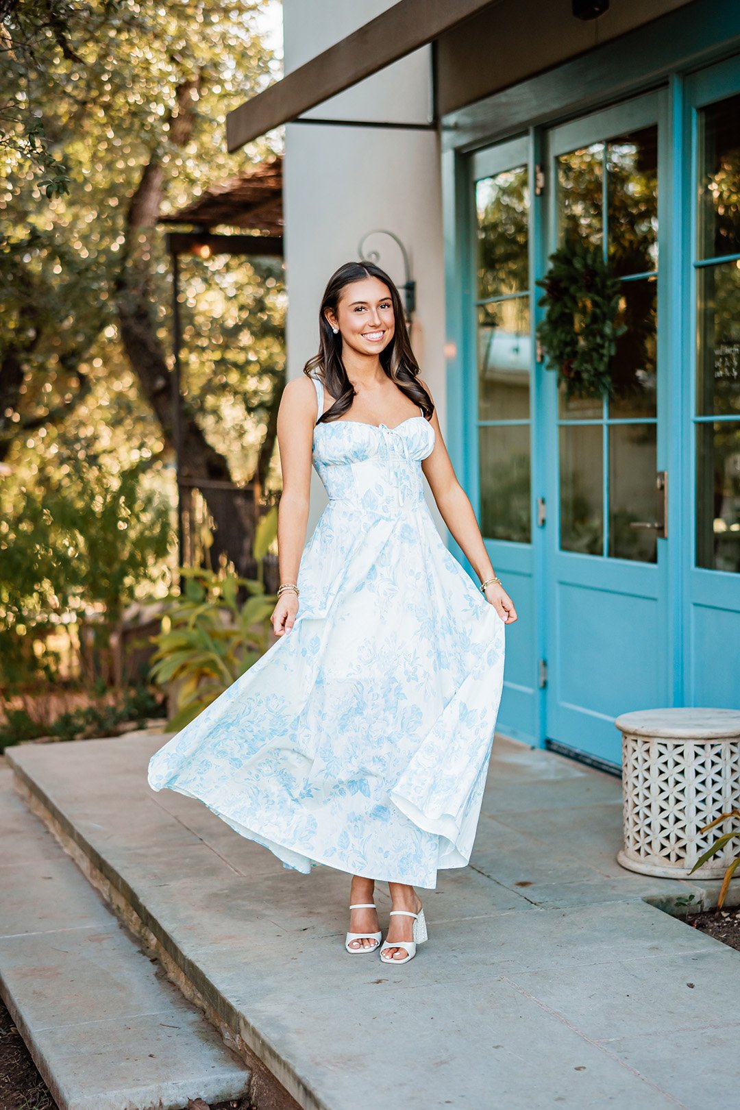A young woman in a light blue floral dress and white high-heeled sandals standing outside a house with blue doors, smiling.