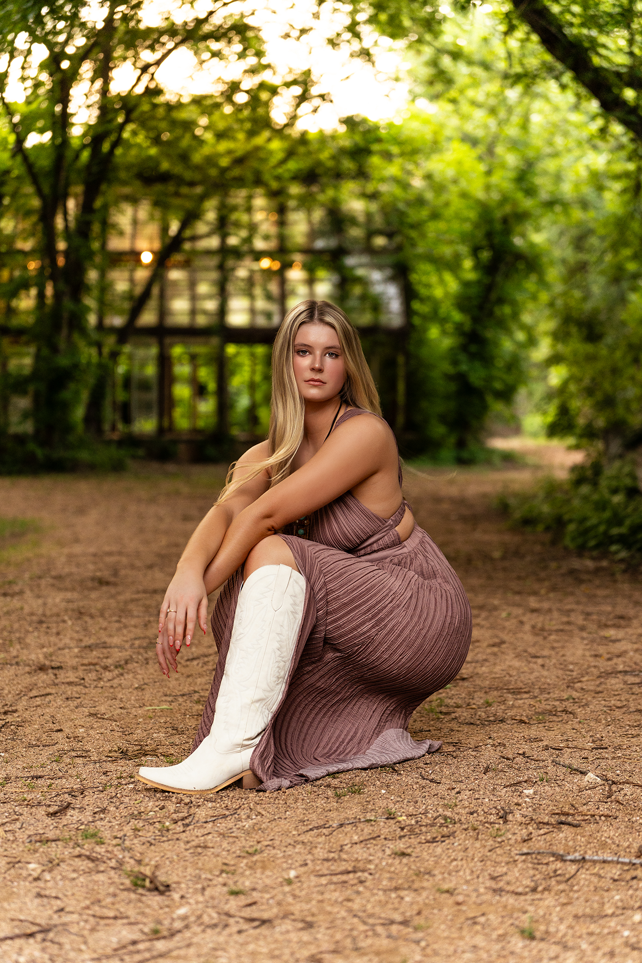 A young woman with long blonde hair sitting outdoors on a dirt path surrounded by green trees during daylight. She is wearing a purple, pleated dress with cutout sides and white cowboy boots.