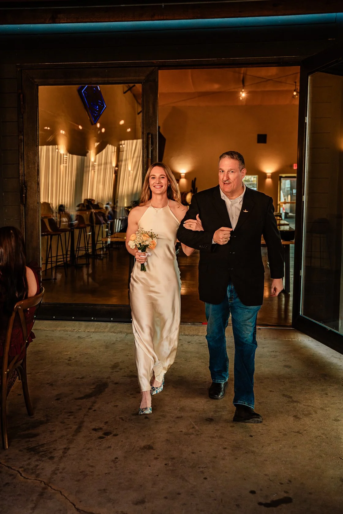 A young woman in a satin dress holding a bouquet walks through a door leading into a warmly lit indoor space, accompanied by an older man in a dark blazer and jeans. There are guests seated at tables inside, and the setting suggests a celebration or 