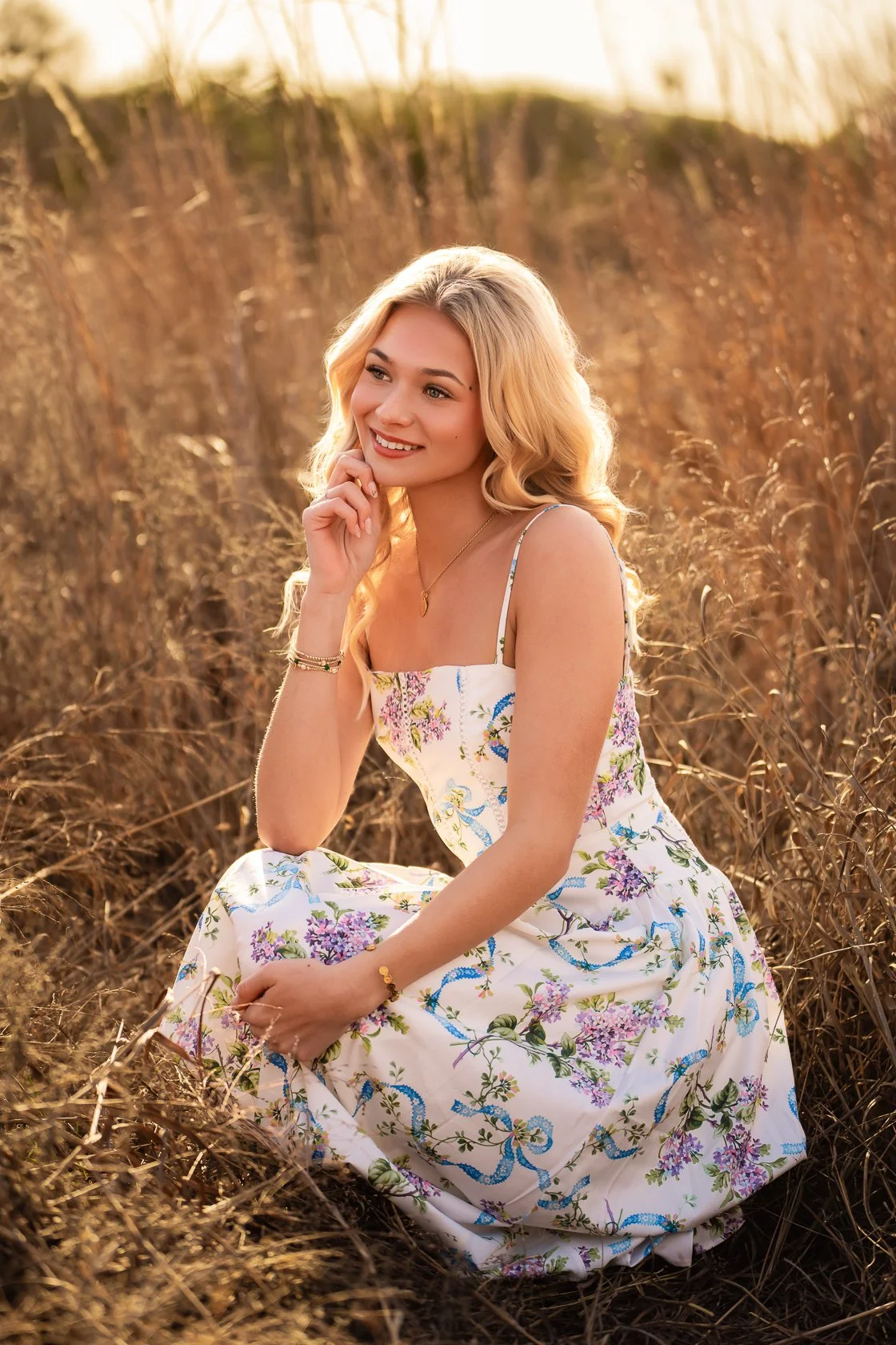A young woman with long blonde hair in a white floral dress sitting in a field of tall grass during sunset, smiling and looking off to the side.