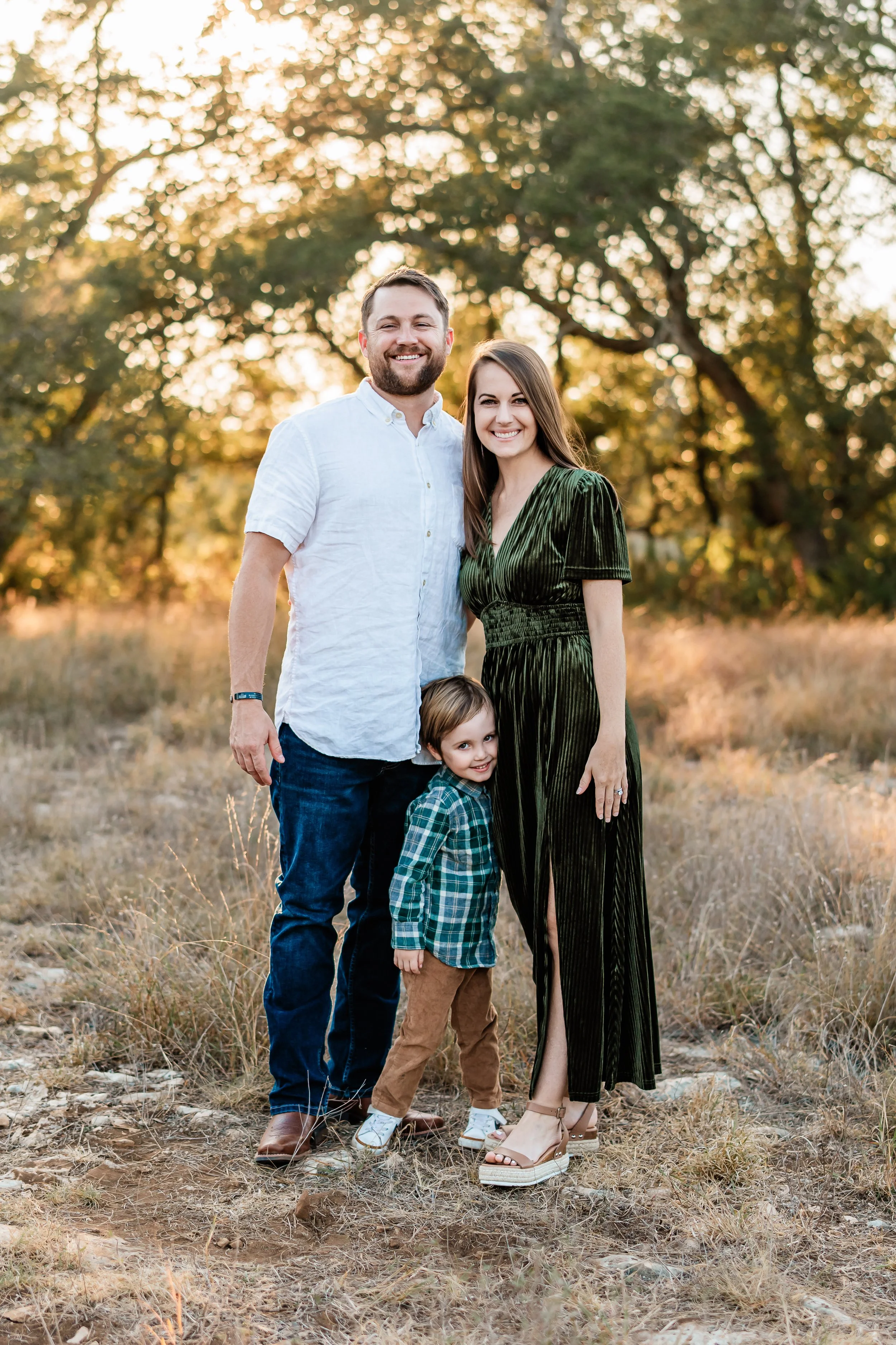 Family of three, a man, a woman, and a young boy, standing outdoors in a grassy field with trees in the background during sunset. The man is wearing a white shirt, the woman in a dark green dress, and the boy in a green plaid shirt and tan pants, all