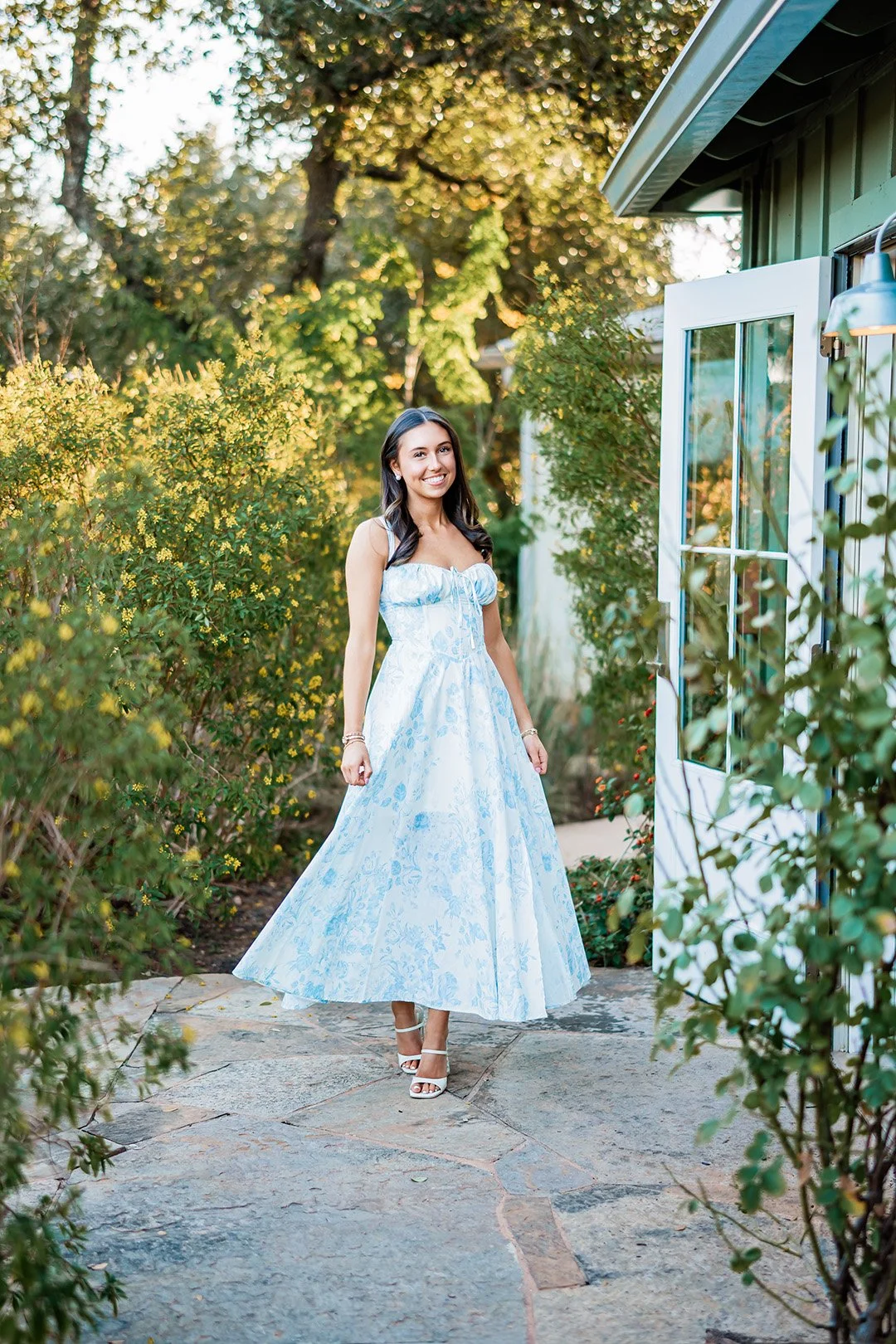 A young woman in a long, light blue patterned dress and white heels walking outside in a garden area with greenery and a stone pathway.