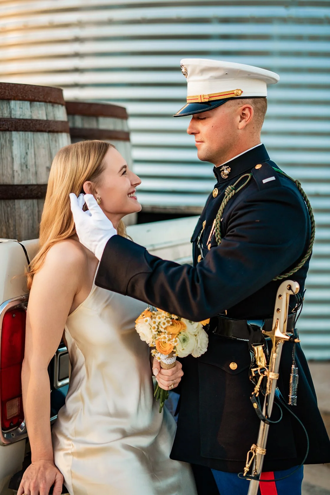 A couple at a wedding, a woman in a white dress holding a bouquet of flowers, smiling at a man in a military uniform, who is touching her face gently.