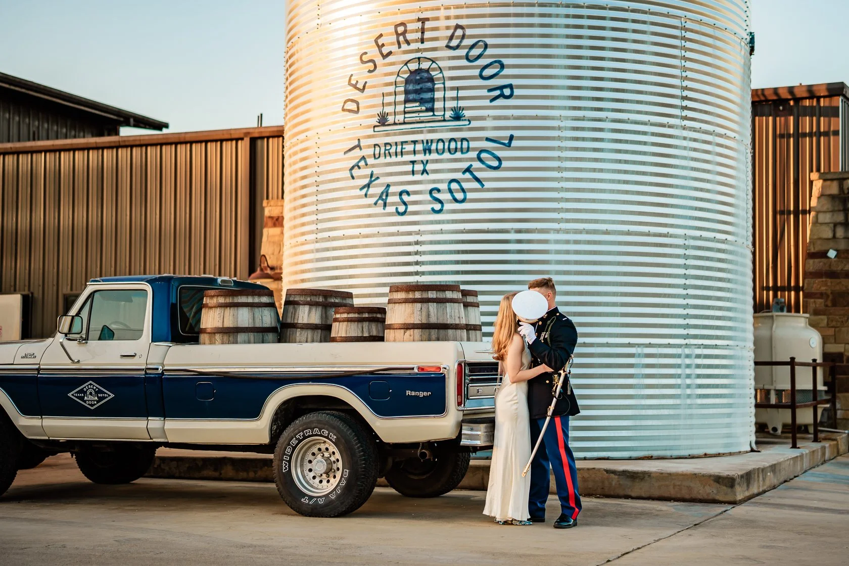 A couple in wedding attire kissing in front of a vintage pickup truck with barrels in the bed, near a large water tower with text 'Desert Door Texas Sotto' and a logo, in an industrial setting.